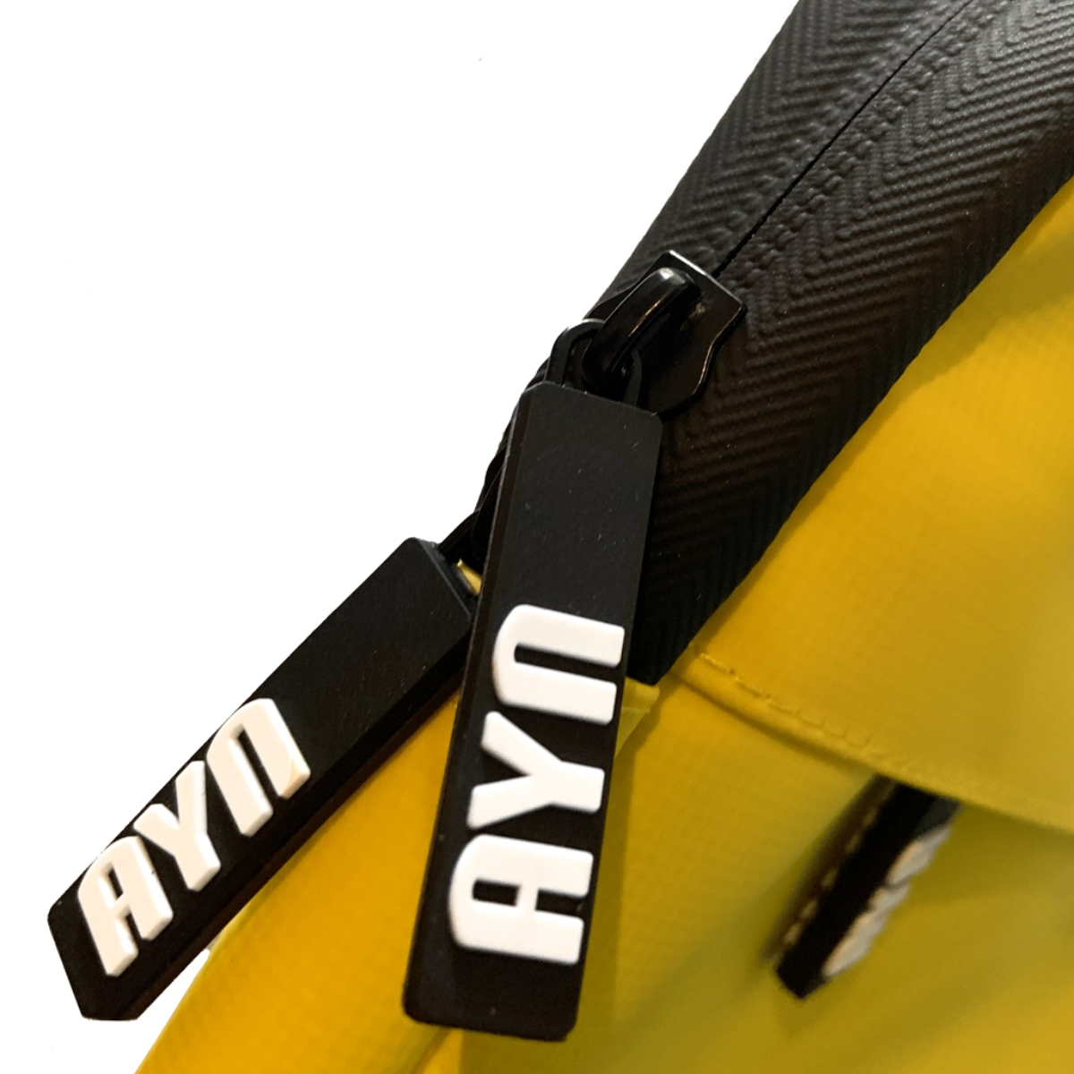 Primer plano de una bolsa amarilla con cremalleras negras. Las cremalleras tienen el texto 'AYN' en blanco.