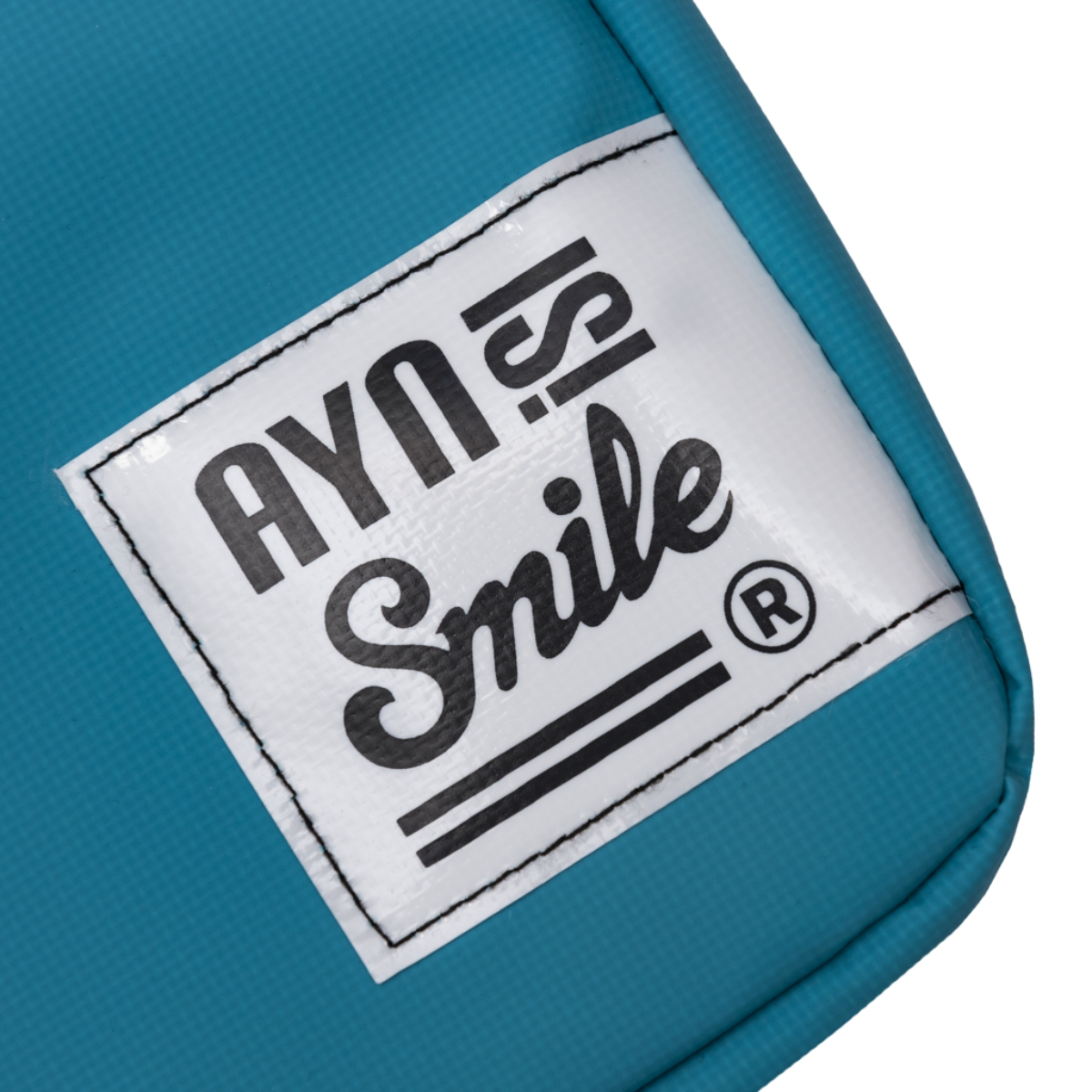 Primer plano de una etiqueta blanca en una bolsa turquesa con el logo 'AYN Smile'.