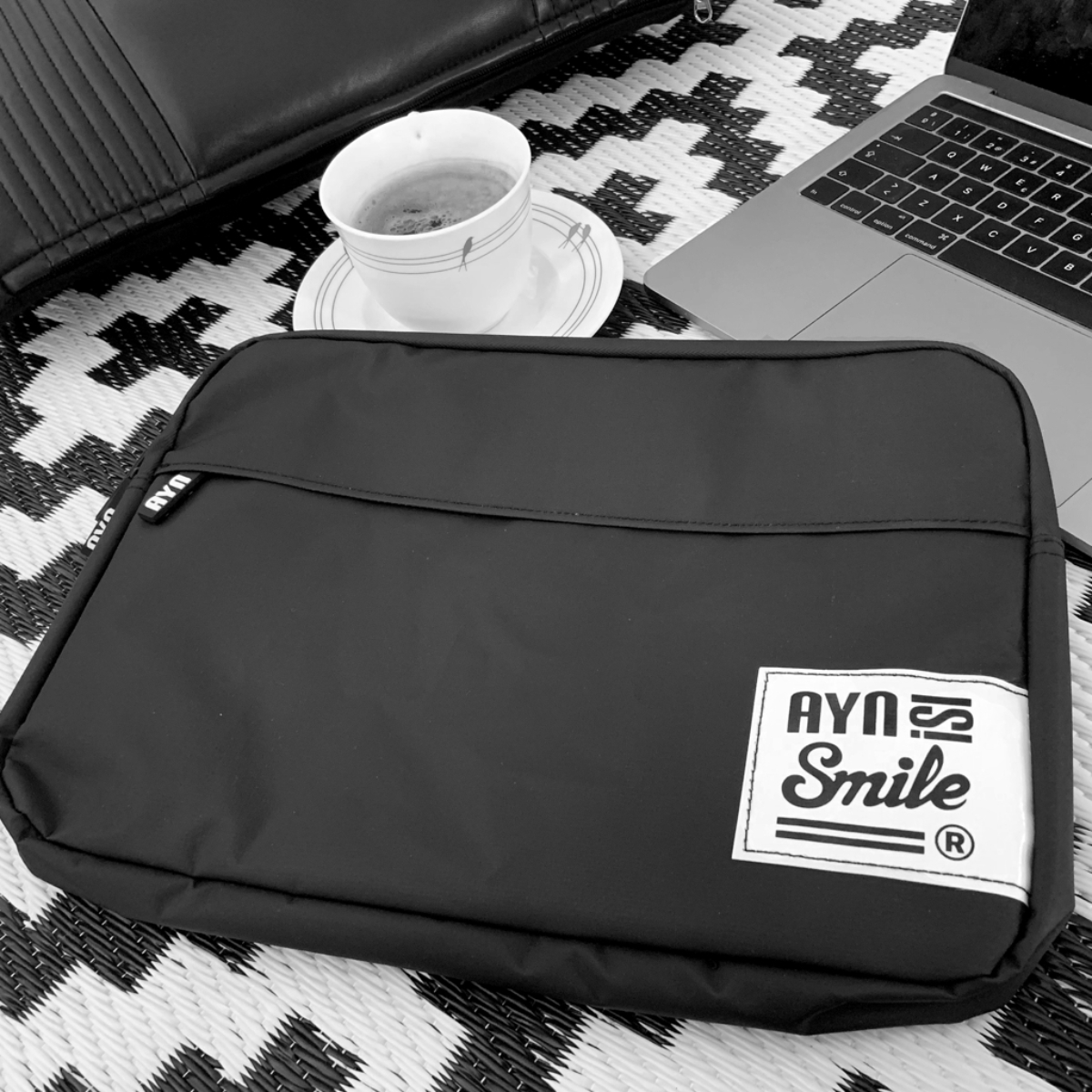 Una custodia per laptop nera con un'etichetta bianca 'AYN Smile', un laptop e una tazza di caffè.