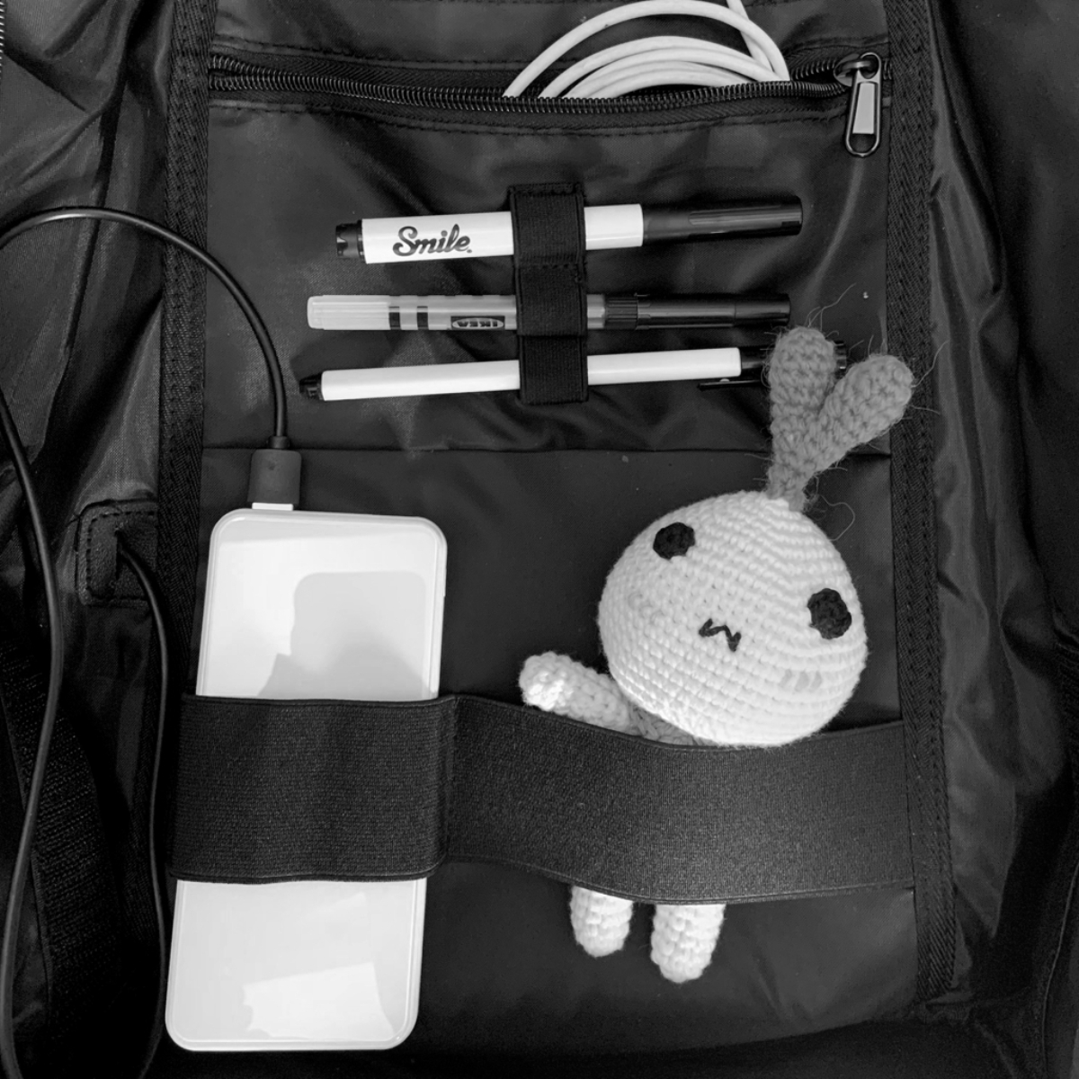 Imagen en blanco y negro del interior de una mochila. Contiene un banco de energía, bolígrafos y un conejo de ganchillo.