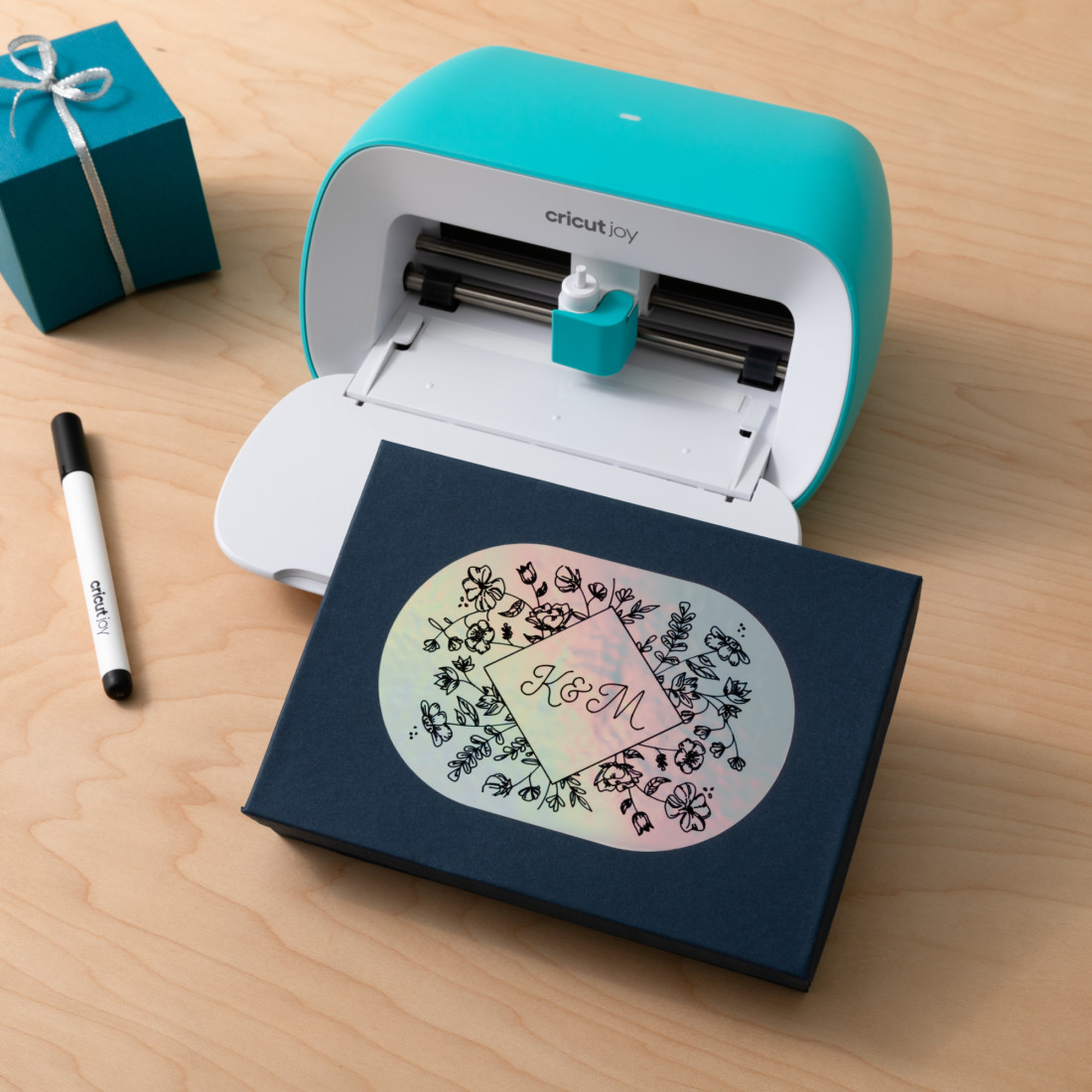 Eine türkisfarbene Cricut Joy Maschine mit einer Geschenkbox und einem schwarzen Stift auf einem Holztisch.