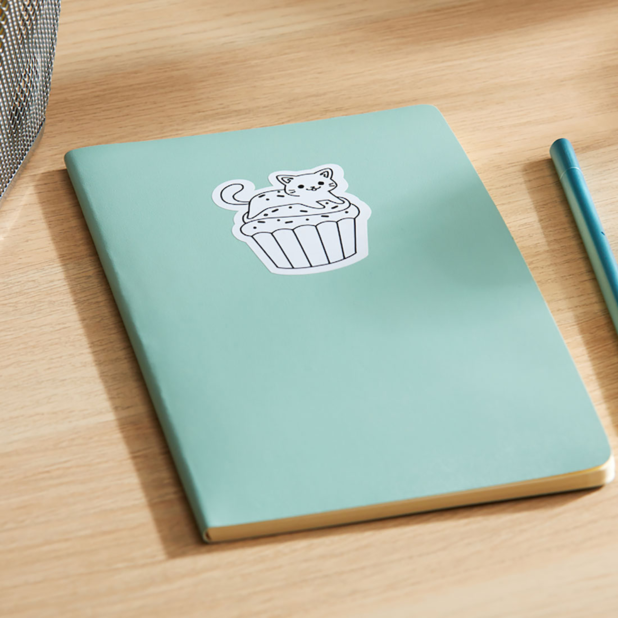 Ein Notizbuch mit einem Katzen-Cupcake-Aufkleber und einem Stift auf einer Holzoberfläche.