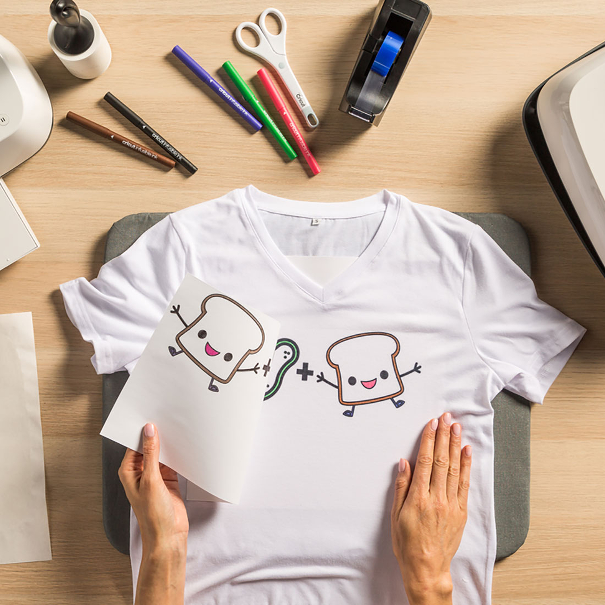 Hände halten ein Papier mit Toast-Design. Ein weißes T-Shirt mit gleichem Design liegt auf einem grauen Pad.