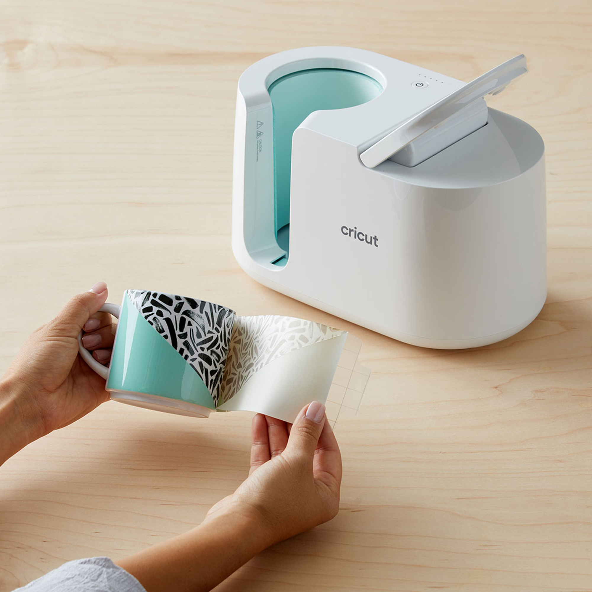 Eine Cricut-Tassenpresse mit einer dekorierten Tasse. Hände ziehen einen gemusterten Aufkleber ab.