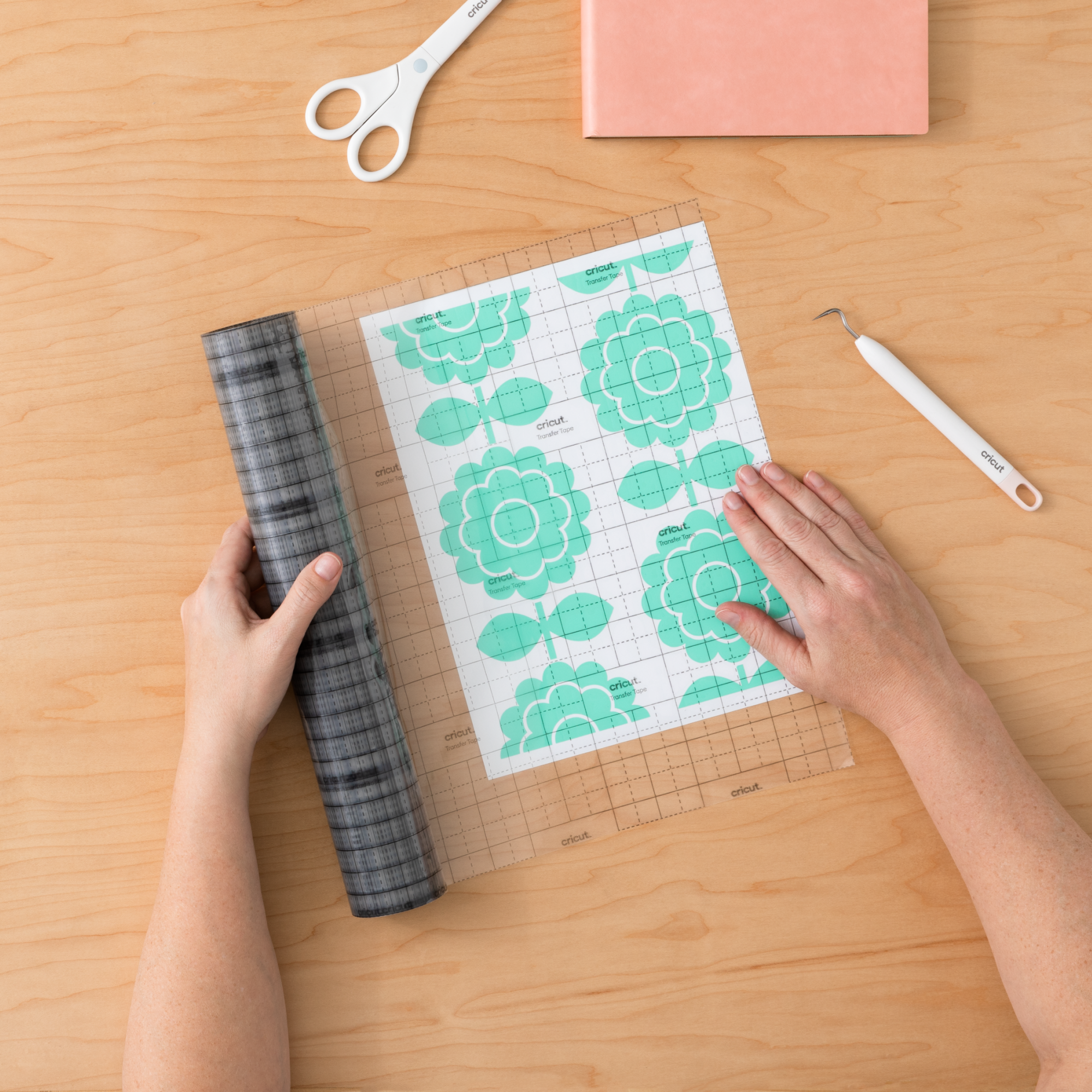 Hände, die ein Werkzeug verwenden, um ein Blumendesign auf einer transparenten Schneidematte mit einer karierten Rolle und Cricut-Zubehör anzubringen.
