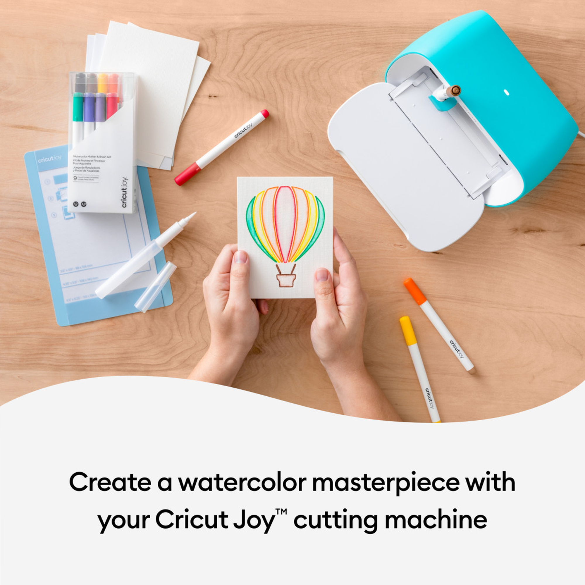 Hände halten eine Aquarellmalerei eines Heißluftballons, Cricut Joy Maschine in der Nähe.