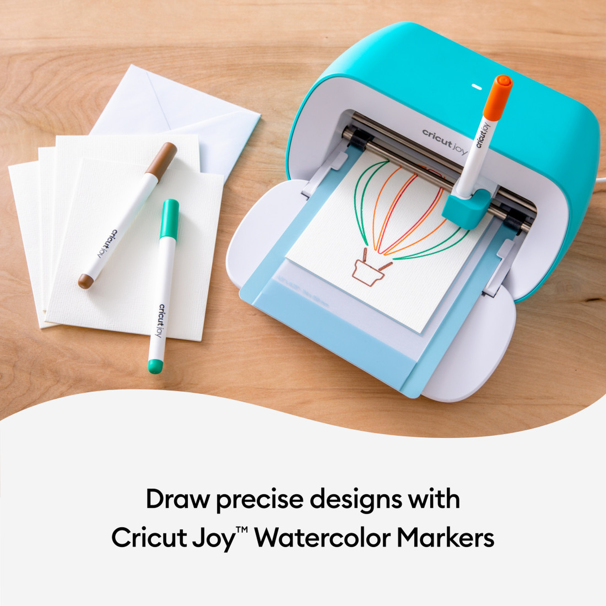 Eine Cricut Joy Maschine, die mit bunten Stiften auf Papier zeichnet. Marker und Papier liegen auf einem Holztisch.