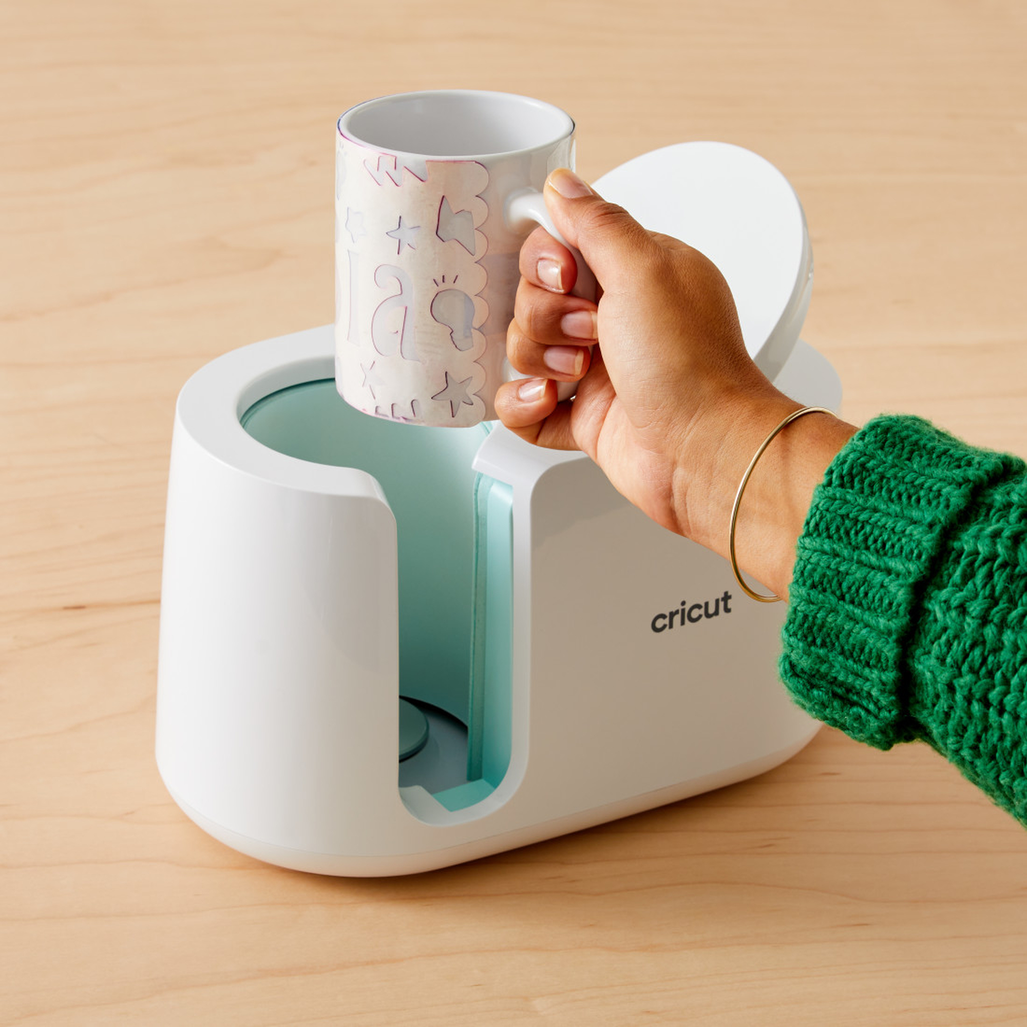Eine Person hält einen weißen Becher mit floralem Design neben einer weißen Cricut-Heißpressmaschine.