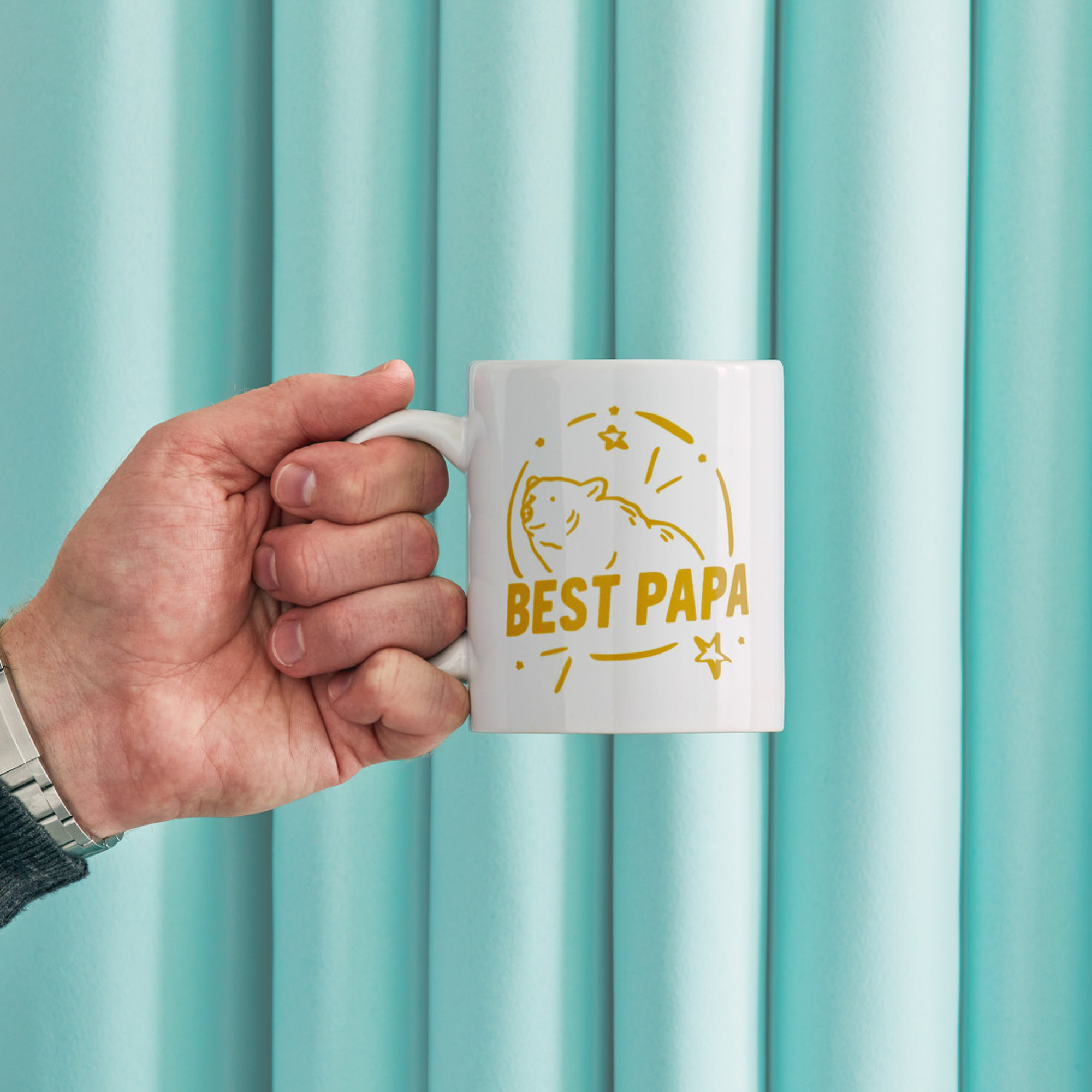 Eine Hand hält einen weißen Becher mit einem goldenen Bären und dem Text „BEST PAPA“.