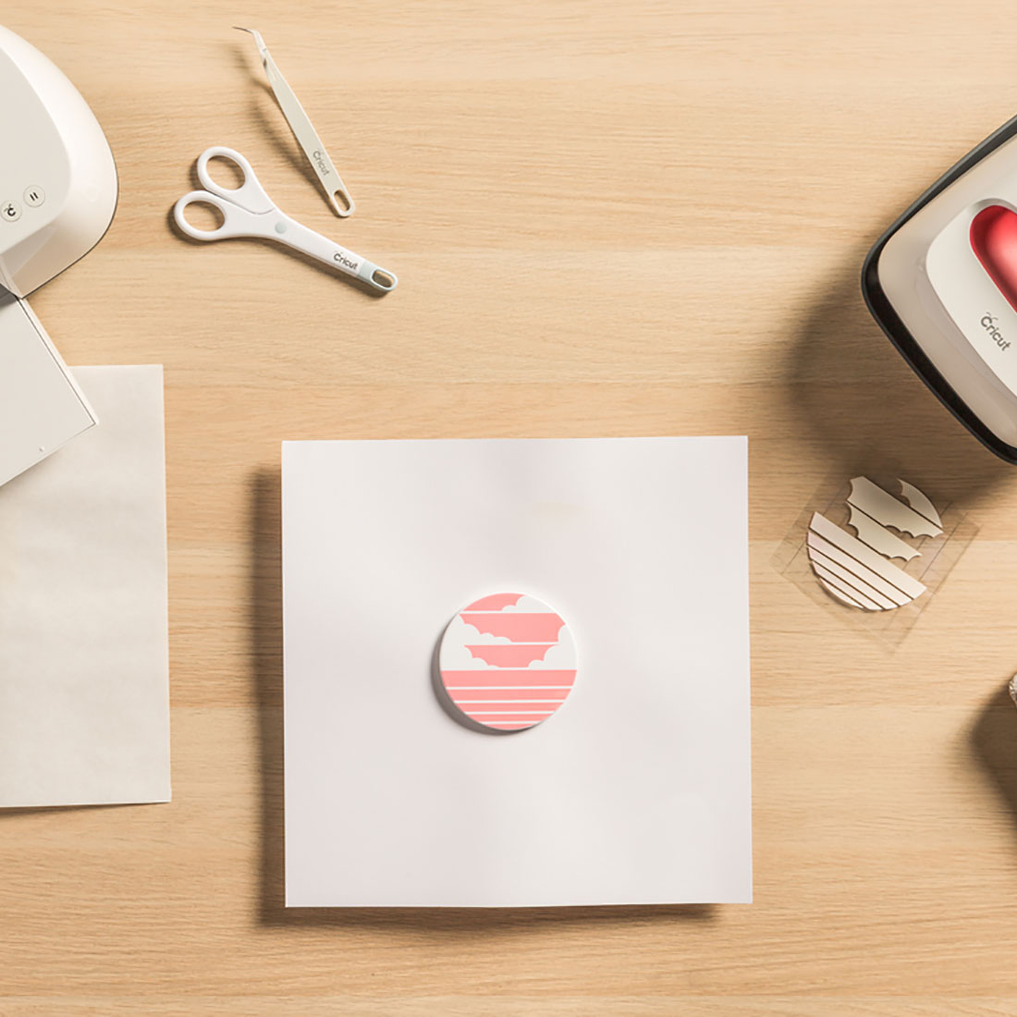 Bastelmaterialien auf einem Holztisch, darunter Schere, eine Cricut-Maschine, Papier und ein dekorativer Aufkleber.