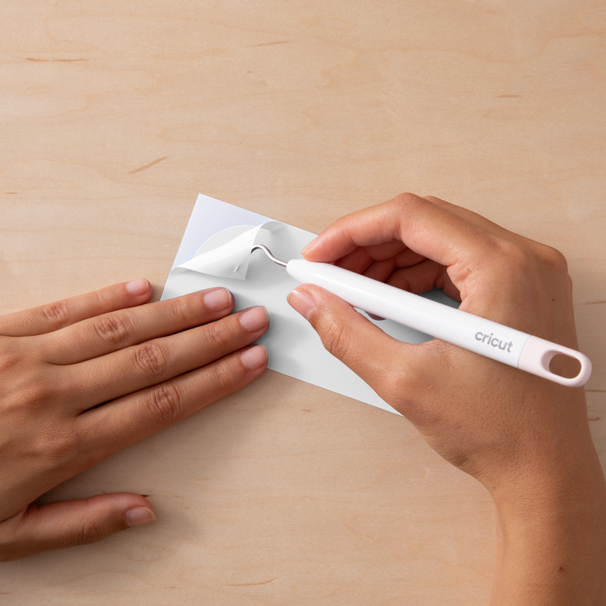 Eine Hand verwendet ein weißes Cricut-Werkzeug, um einen weißen Aufkleber von einem blauen Träger auf einer Holzoberfläche zu entfernen.