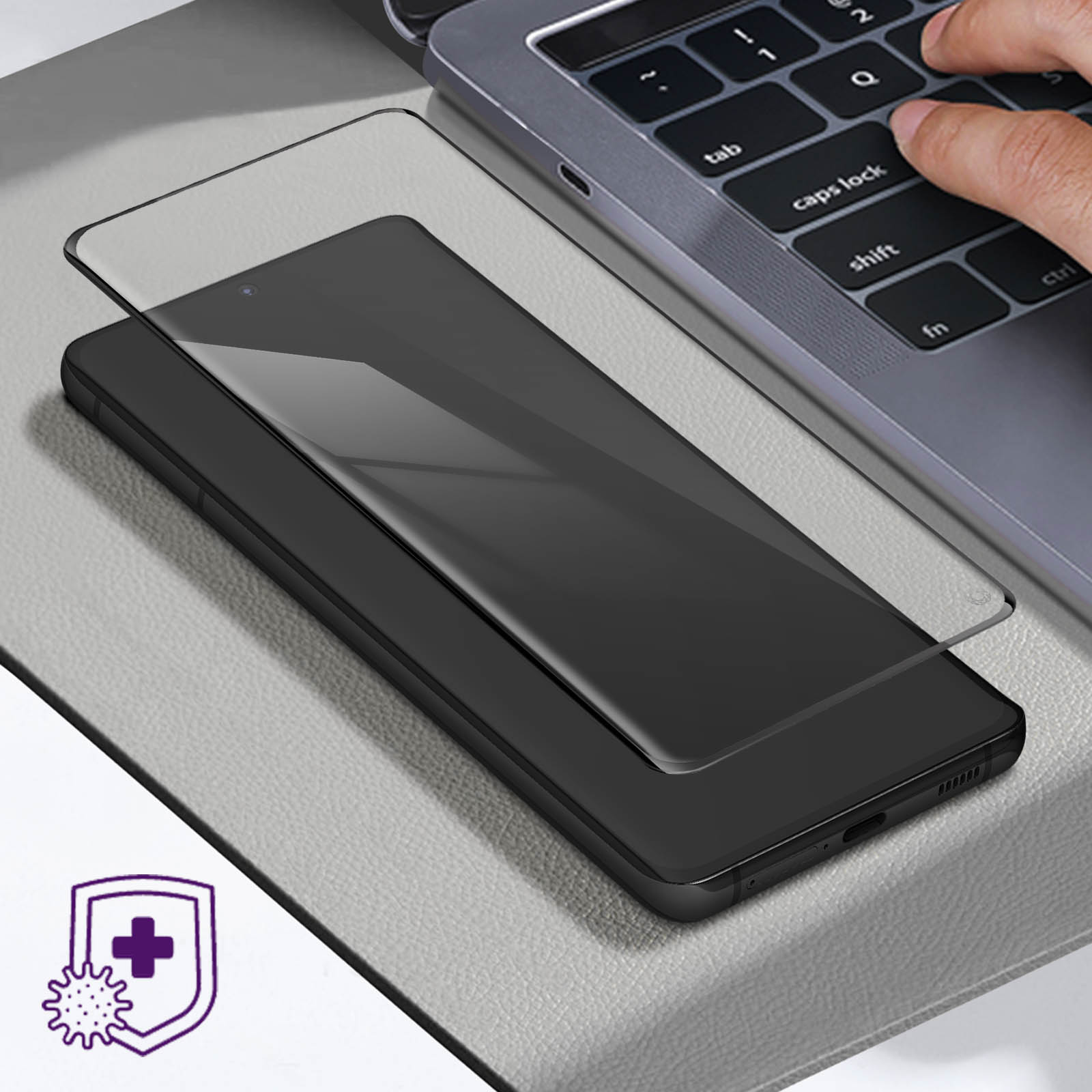Teléfono negro con protector de pantalla sobre una superficie gris, con un portátil detrás.