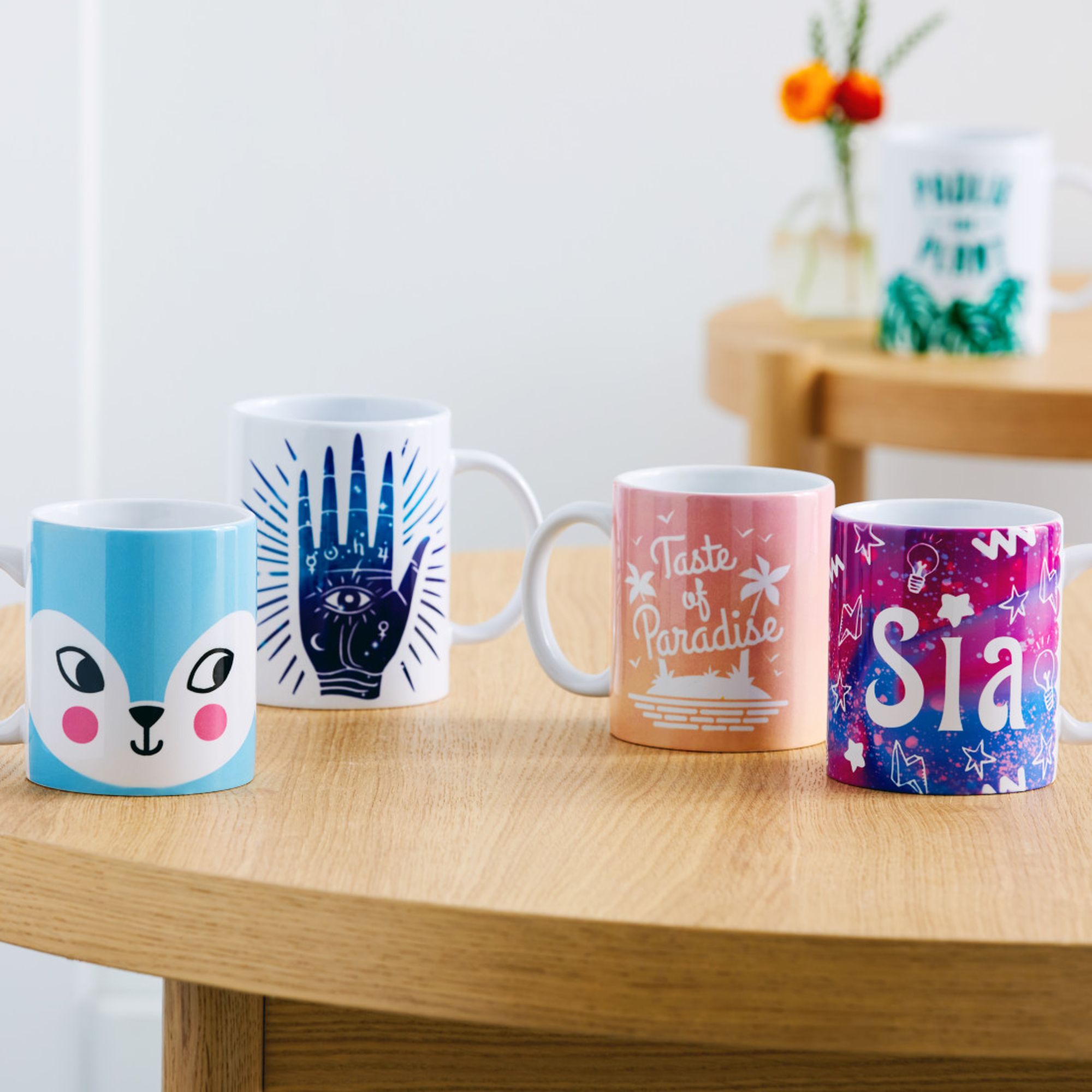 Vier Tassen auf einem Holztisch mit bunten Designs. Eine Vase mit Blumen im Hintergrund.
