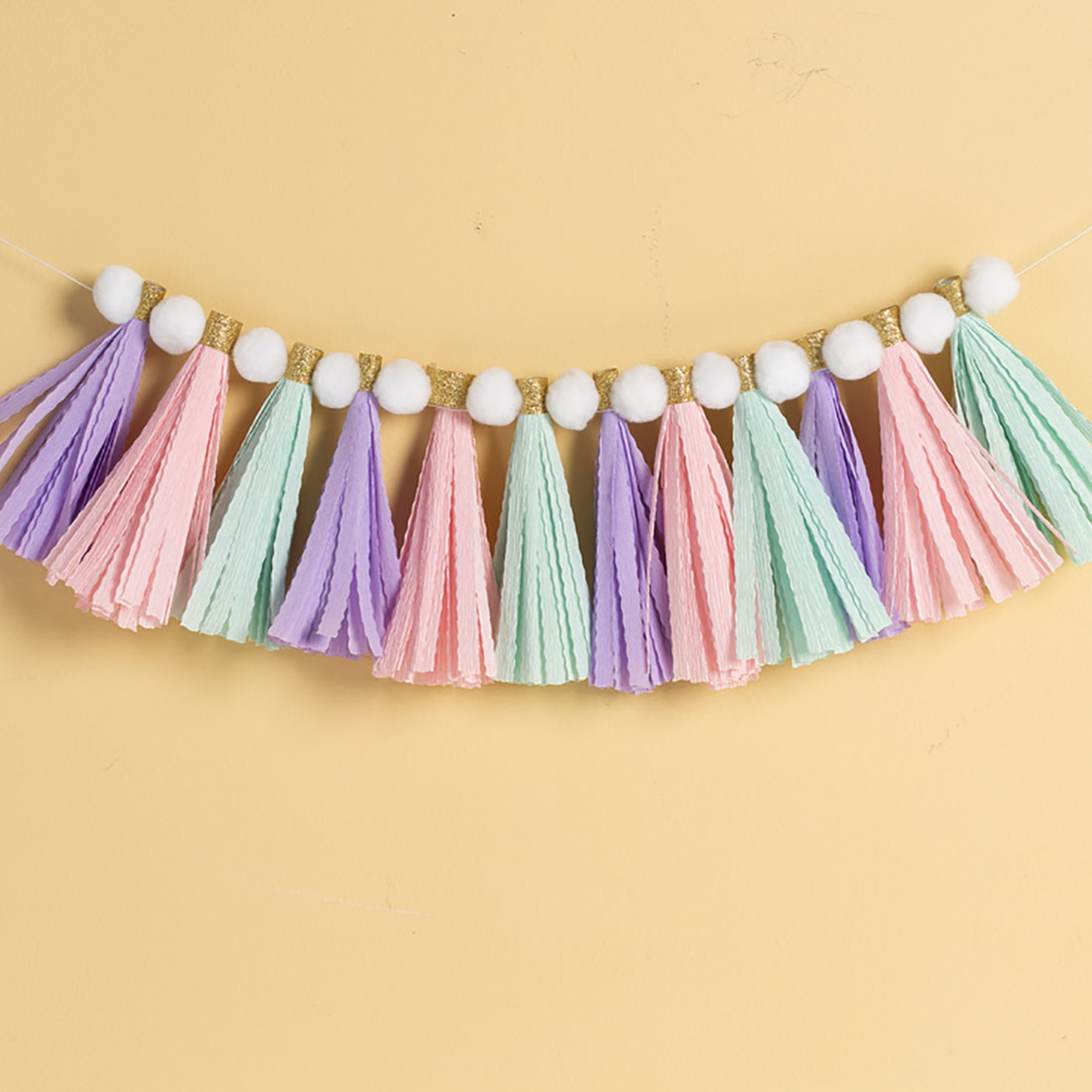Eine bunte Girlande mit pastellfarbenen Quasten und weißen Pompons auf gelbem Hintergrund.