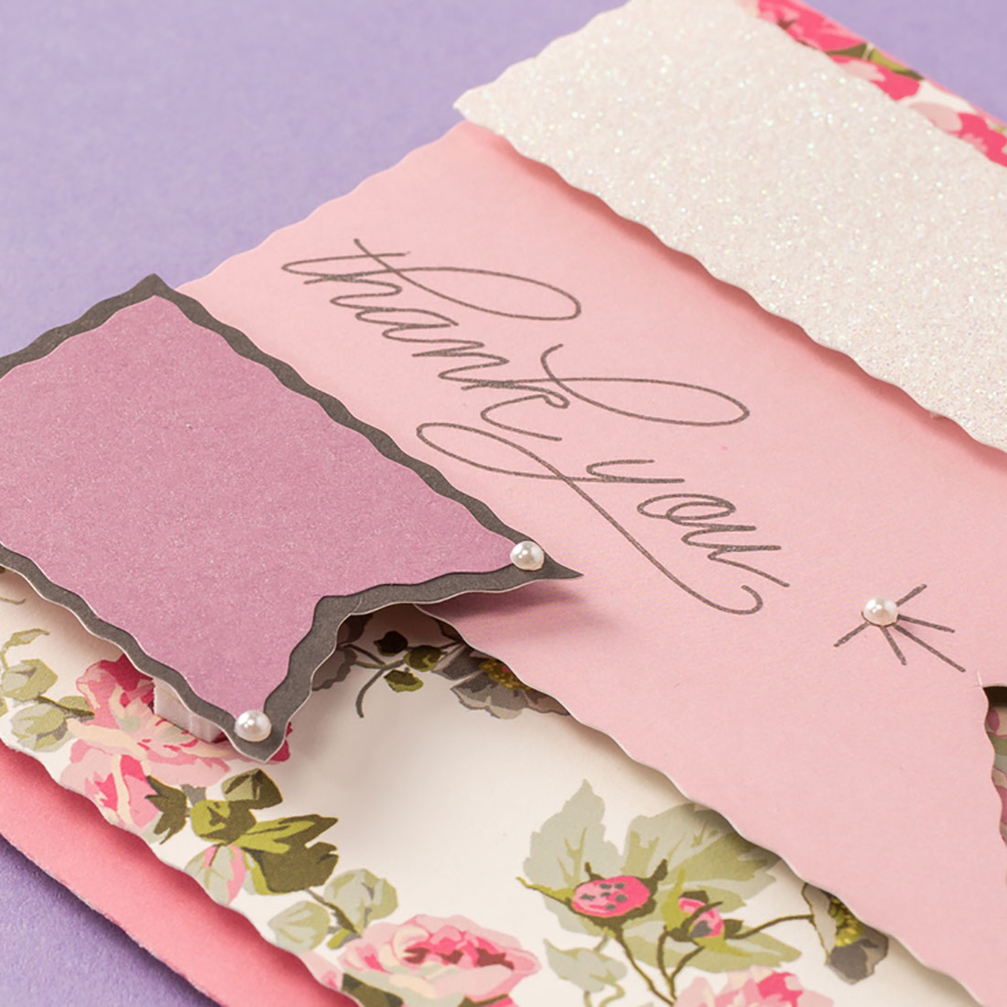 Rosa Karte mit 'Thank You' und Perlen, Papier mit Blumenmuster auf lila Hintergrund.