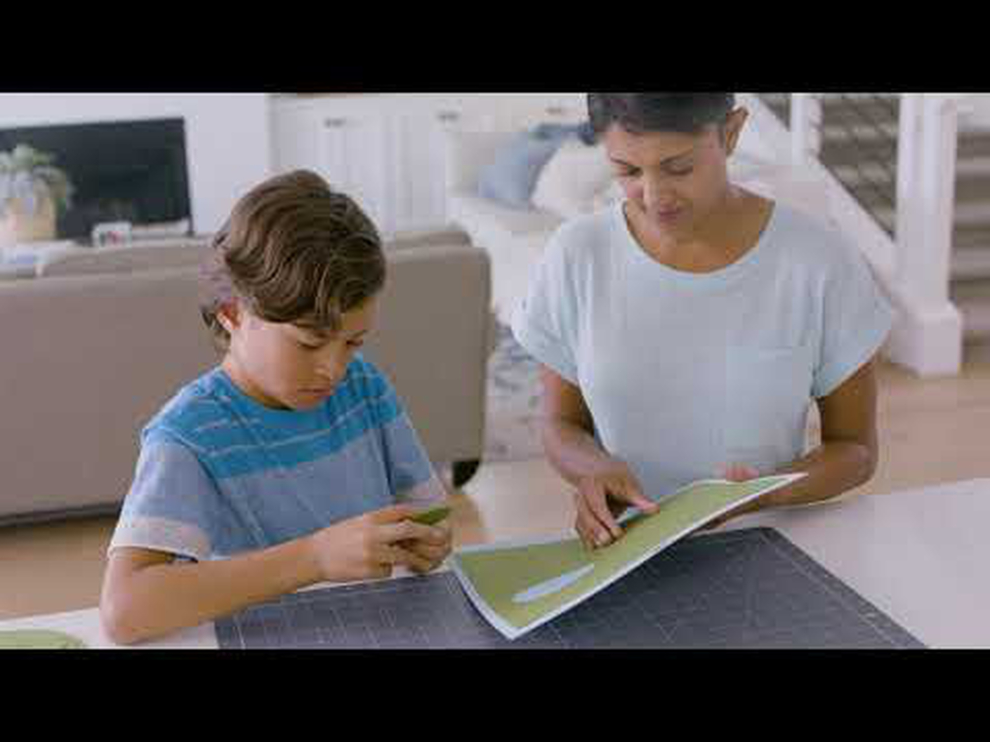 Ein Junge und eine Frau sitzen an einem Tisch mit Papier, im Hintergrund ein Wohnzimmer.
