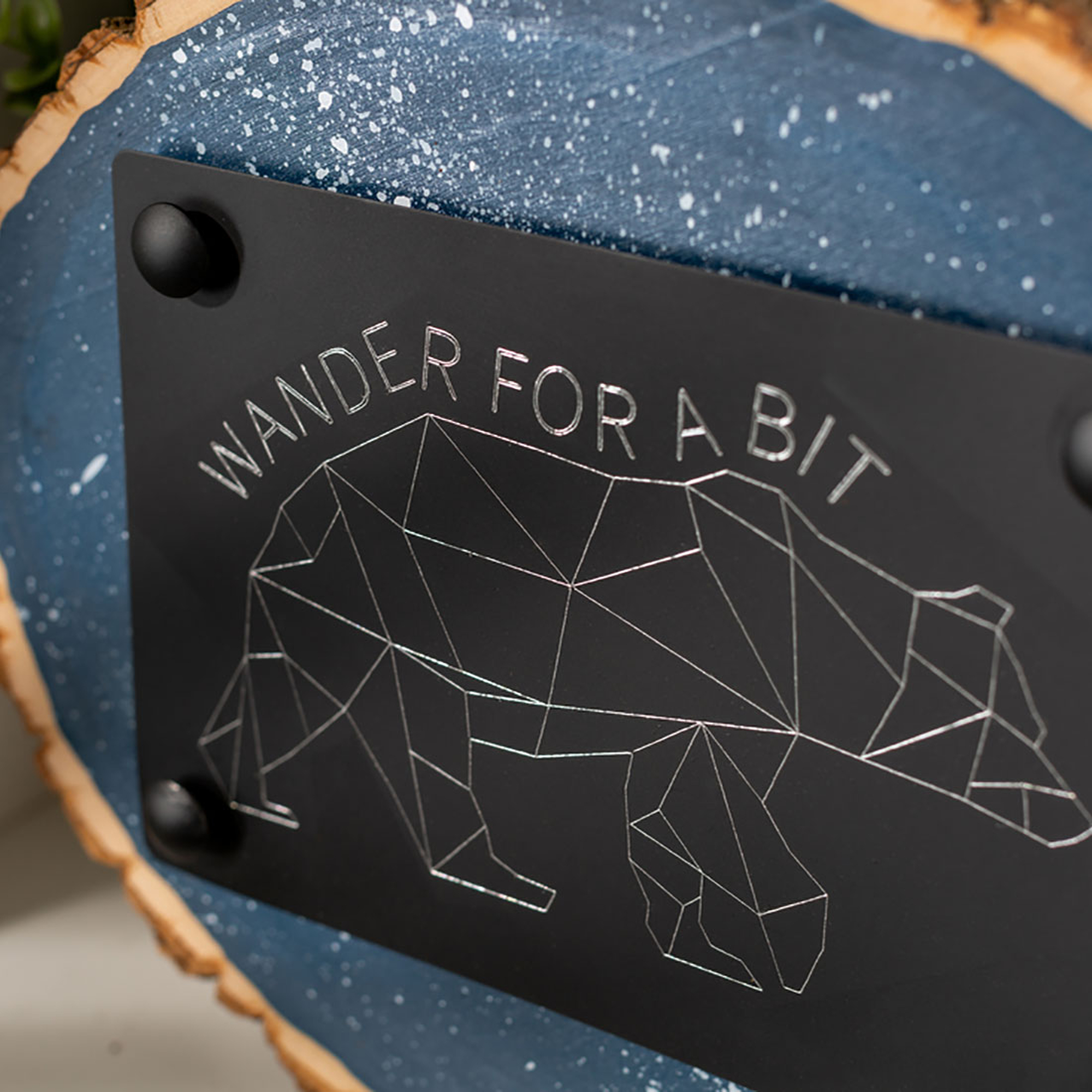 Ein schwarzes Metallschild auf einem blauen Holzstück mit dem Text 'WANDER FOR A BIT' und einer Bärenkontur.