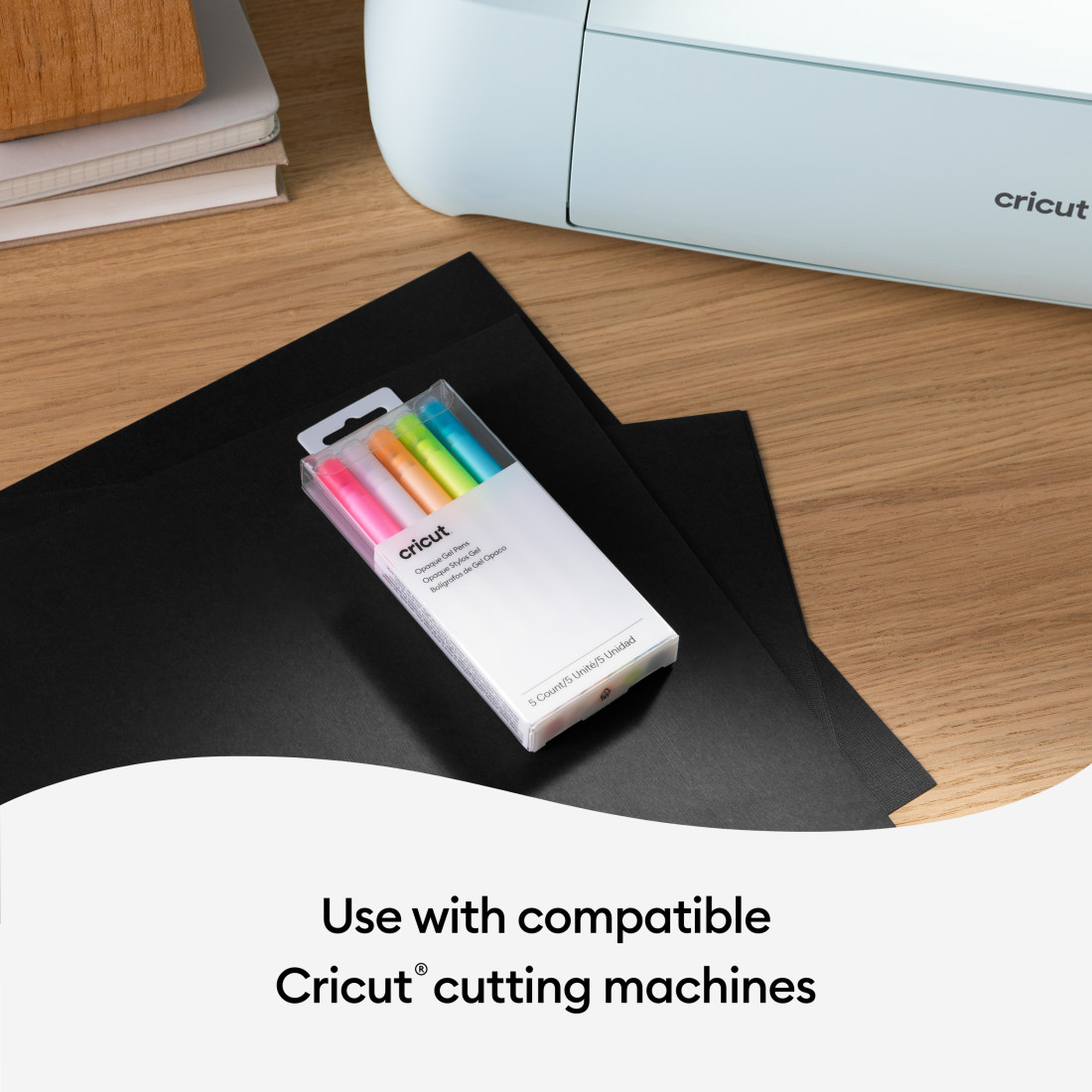 Cricut-Stifte und schwarzes Papier auf einem Tisch. Eine Cricut-Maschine ist im Hintergrund.