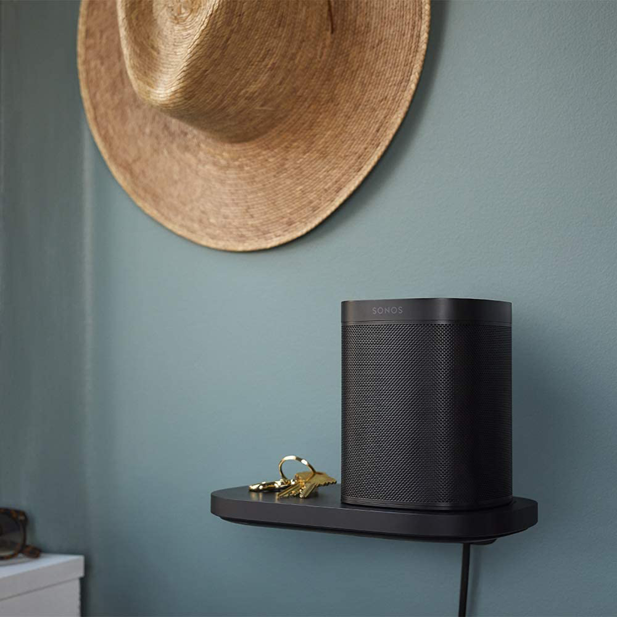 Altavoz negro en un estante negro, llaves y un sombrero en la pared. La pared es verde azulado.