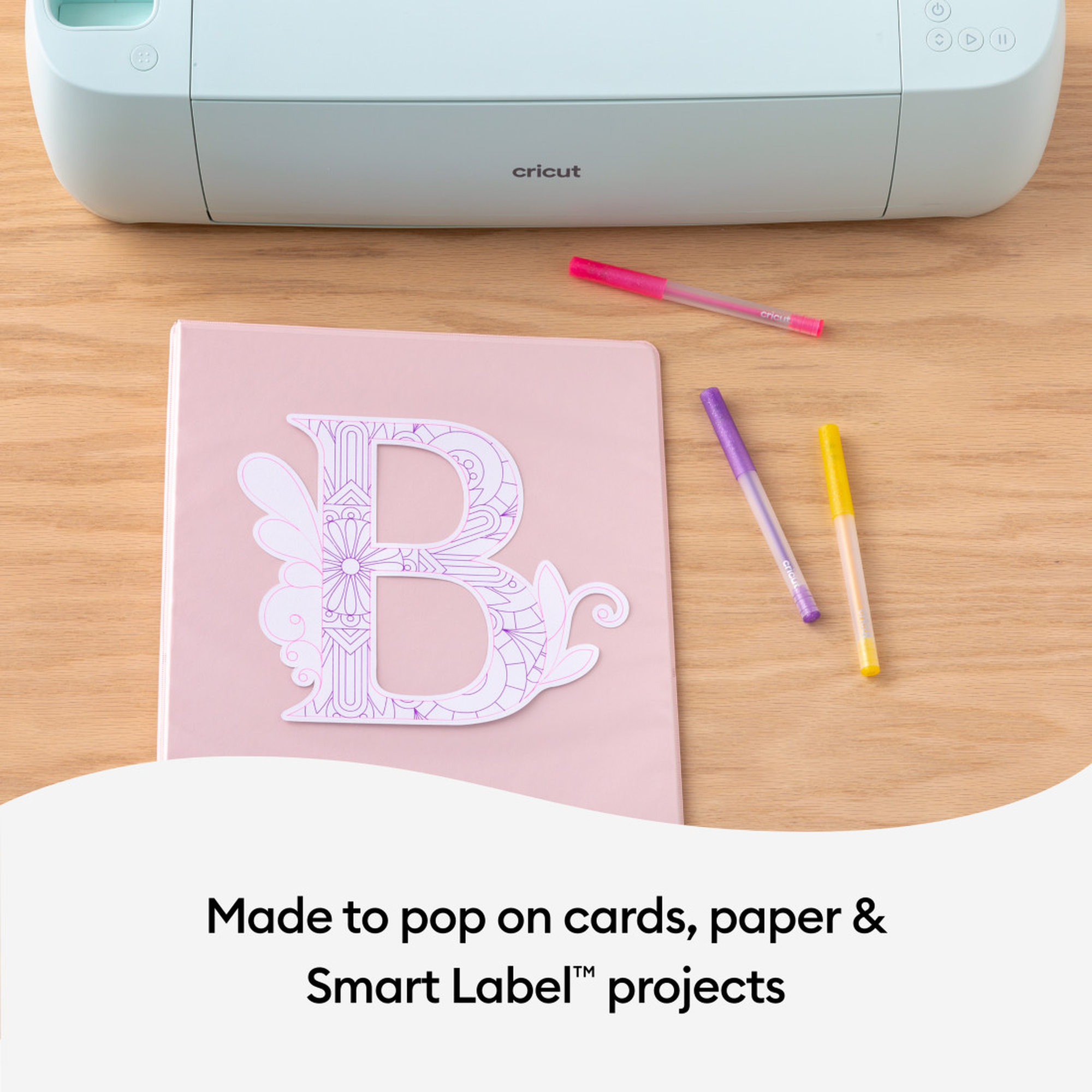 Eine Cricut-Maschine mit einem rosa Notizbuch mit einem ausgeschnittenen Buchstaben 'B', umgeben von Stiften.