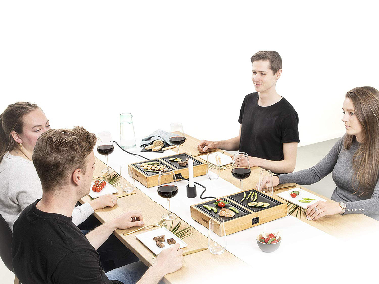 Vier Personen um einen Holztisch mit kleinen Grills und Weingläsern, die gemeinsam Essen zubereiten.