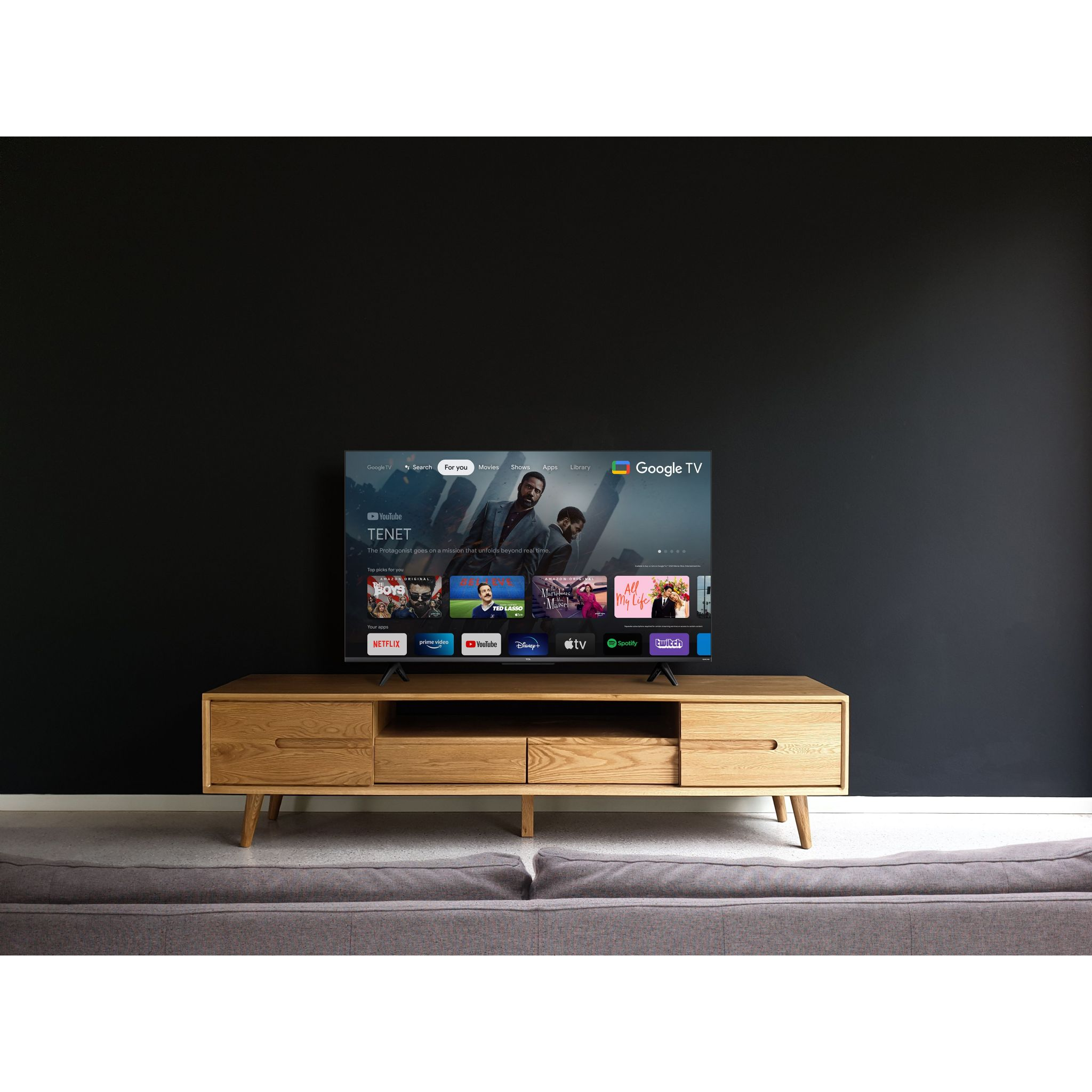 TV en un soporte de madera. Muestra un menú de Google TV y una pared oscura al fondo.
