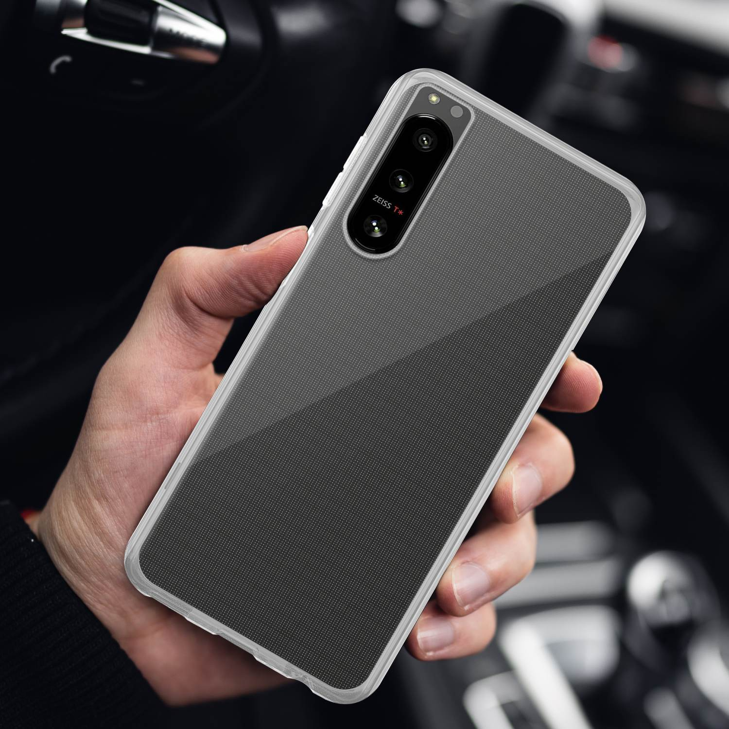 Persona che tiene un telefono con una custodia grigia. Lo sfondo mostra un interno auto.