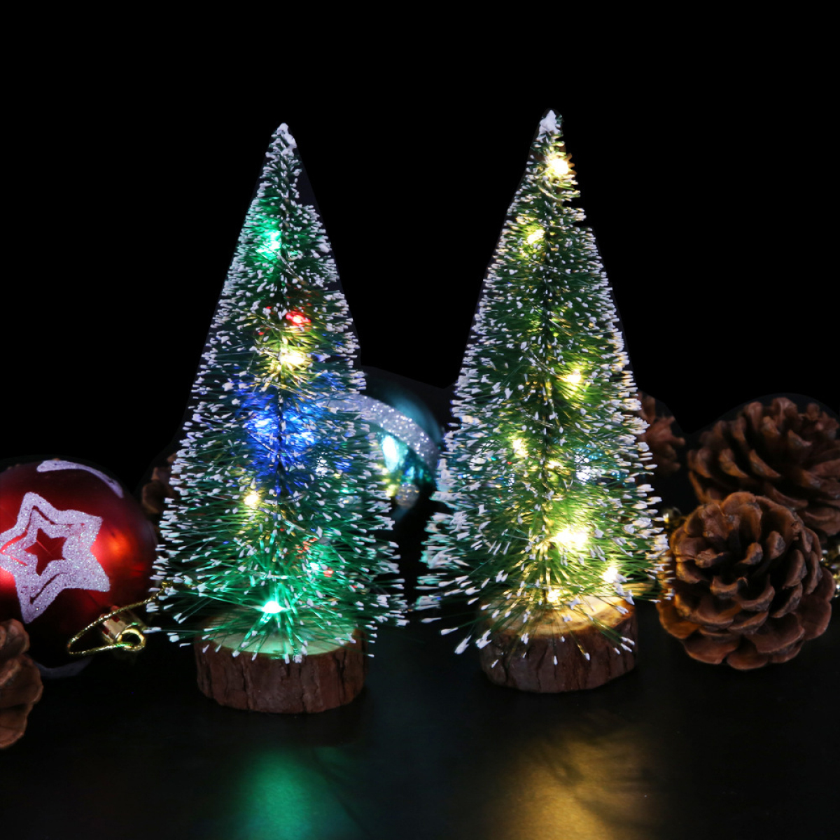 Zwei kleine, beleuchtete Weihnachtsbäume mit bunten Lichtern, umgeben von Tannenzapfen, auf schwarzem Untergrund.