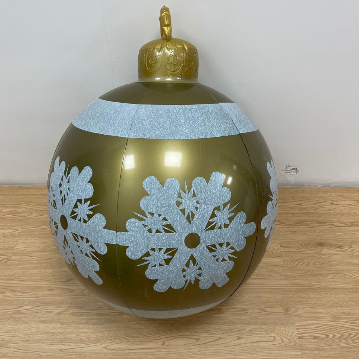Ein großer goldener Weihnachtsschmuck mit weißen Schneeflocken-Designs und einem goldenen Aufsatz, auf Holzboden.