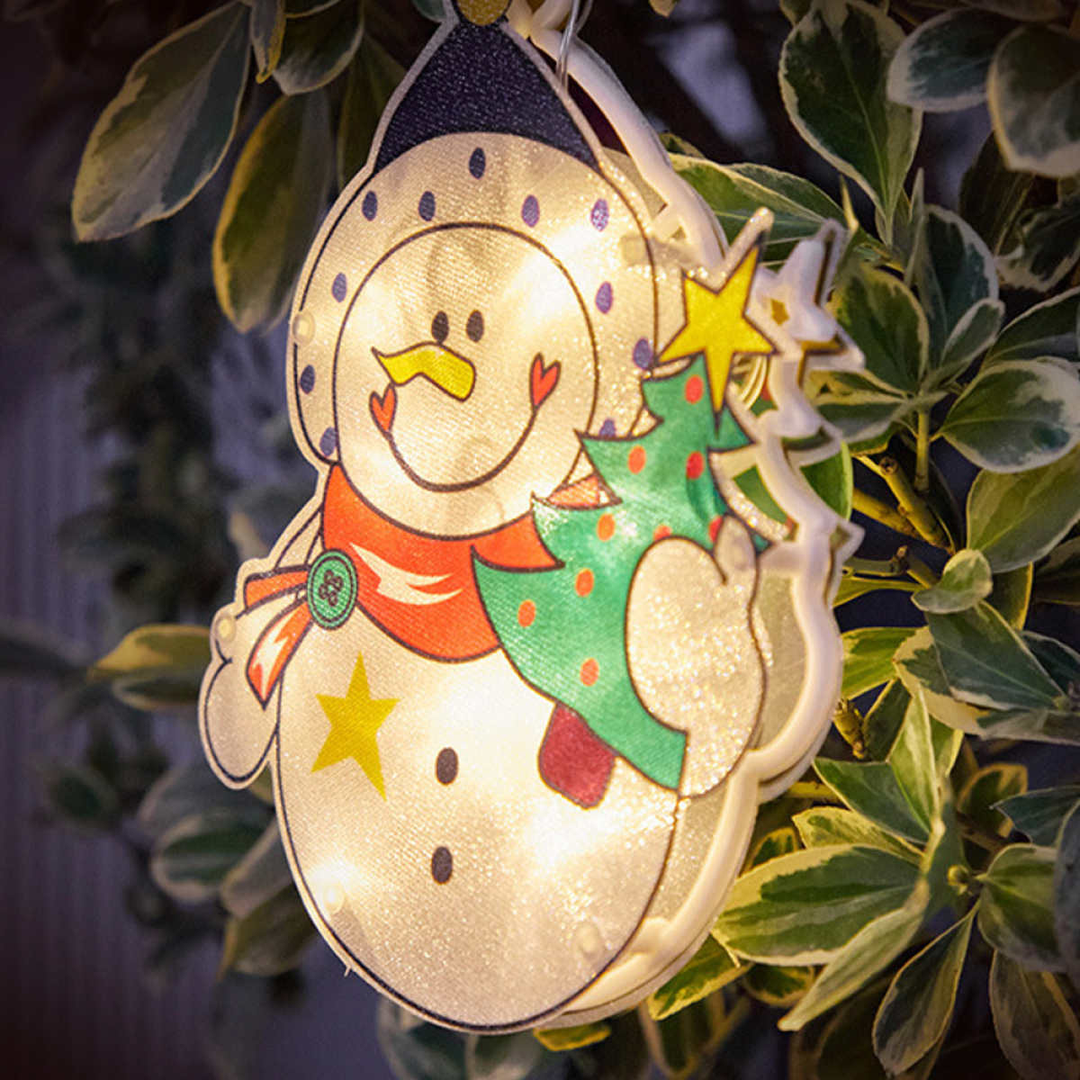 Eine beleuchtete Schneemann-Dekoration mit einem Stern und einem Weihnachtsbaum vor grünen Blättern.