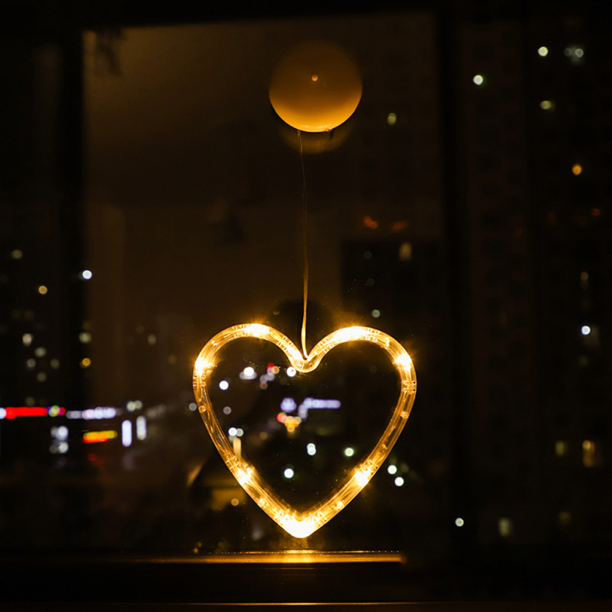Herzförmige beleuchtete Dekoration hängt vor einem Fenster, von außen bei Nacht betrachtet.