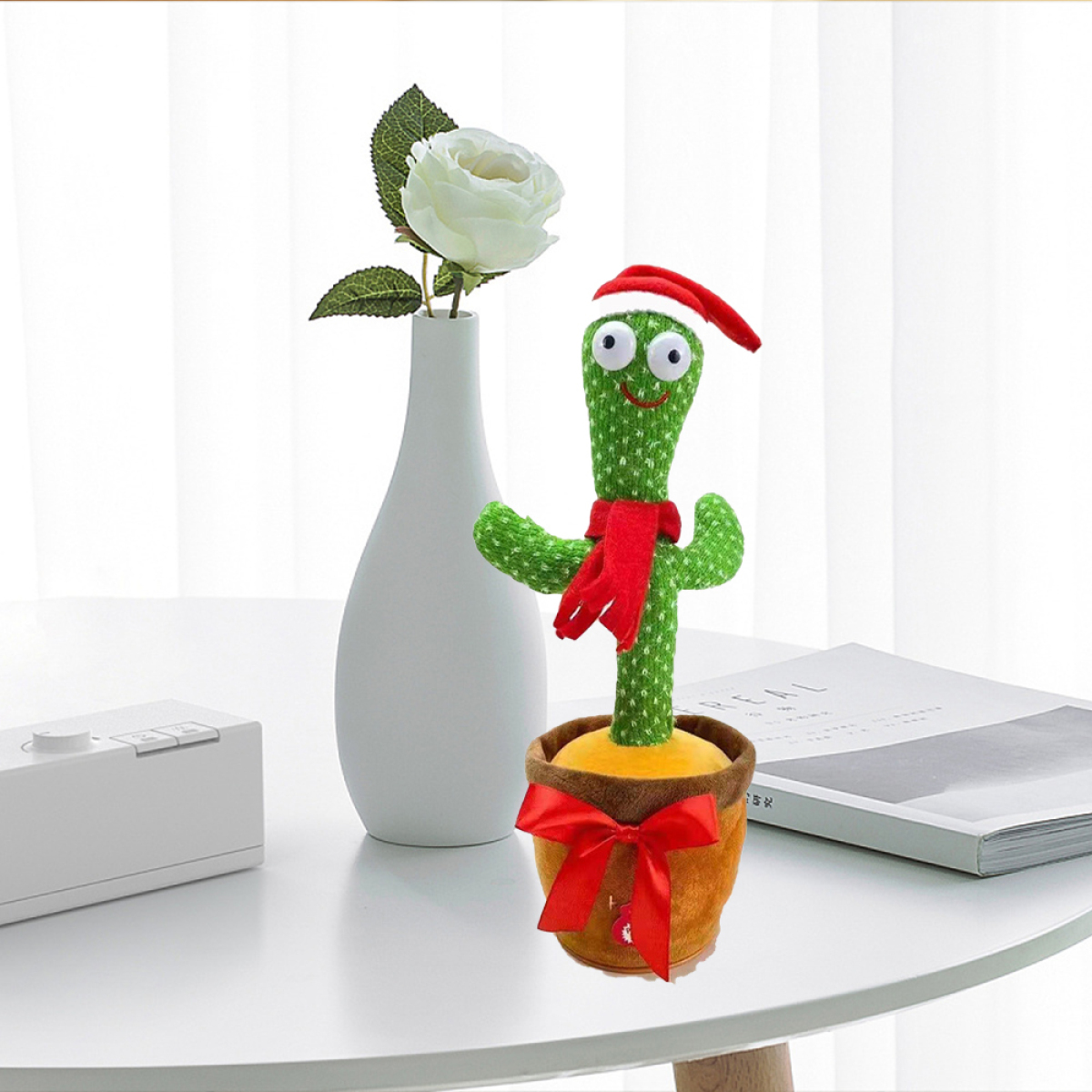 Kaktus-Spielzeug mit Weihnachtsmütze und rotem Schal, in einem Topf mit roter Schleife, neben einer weißen Vase.