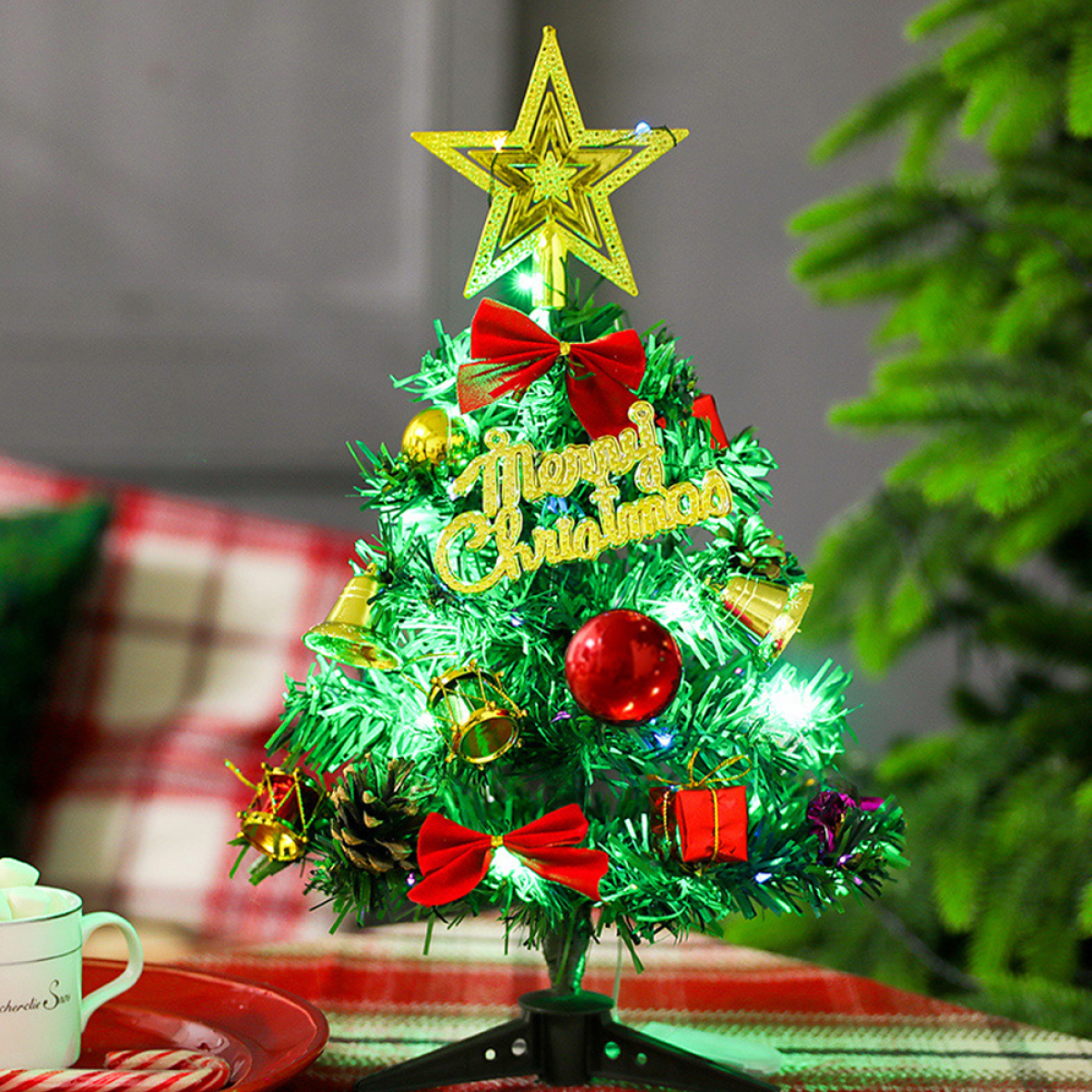 Kleiner Weihnachtsbaum mit Stern, Schleifen, Ornamenten, Glocken und Merry Christmas-Schild auf einem Tisch.