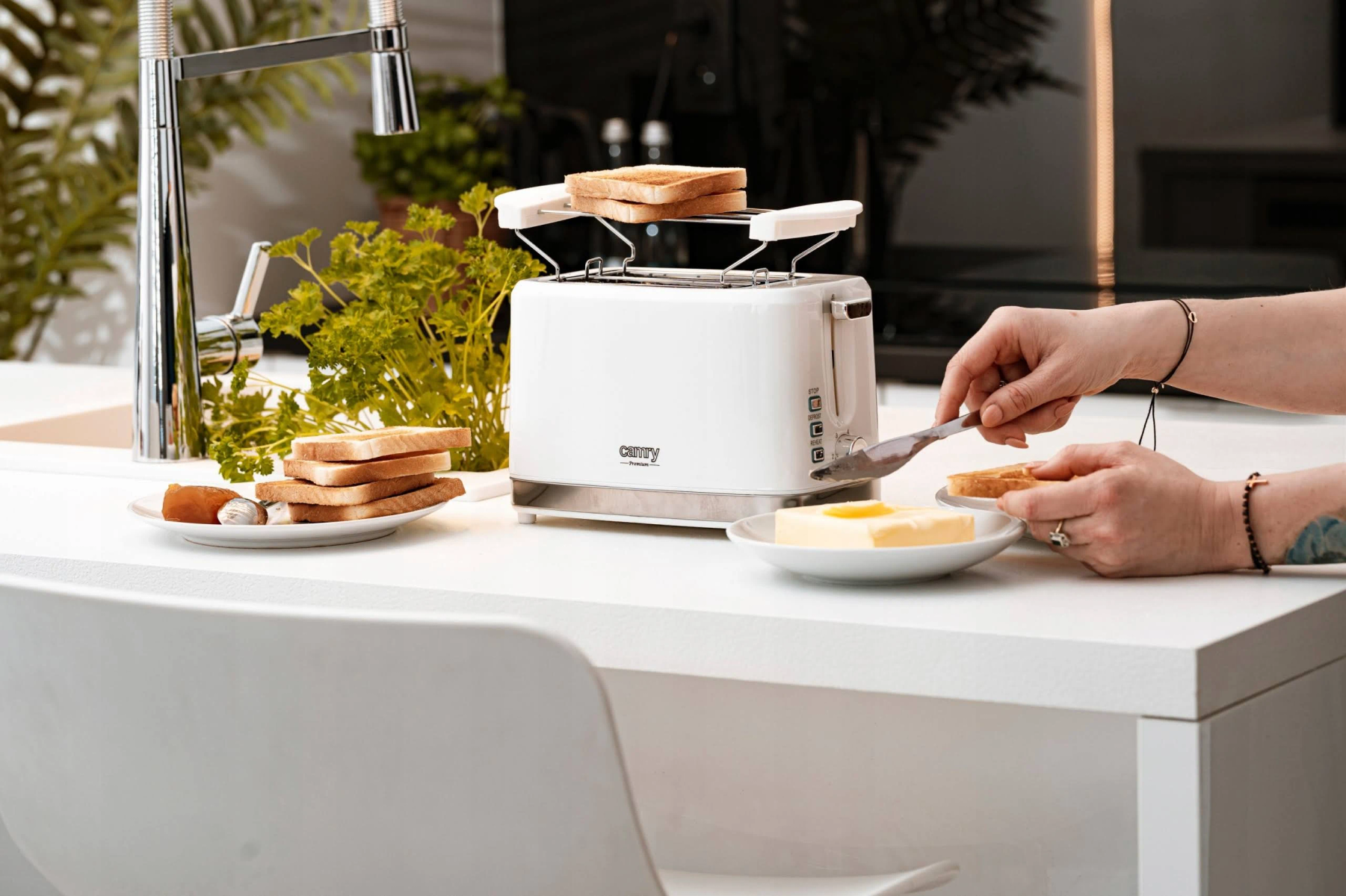 Persona untando mantequilla en tostadas de una tostadora blanca, cerca de platos y plantas.