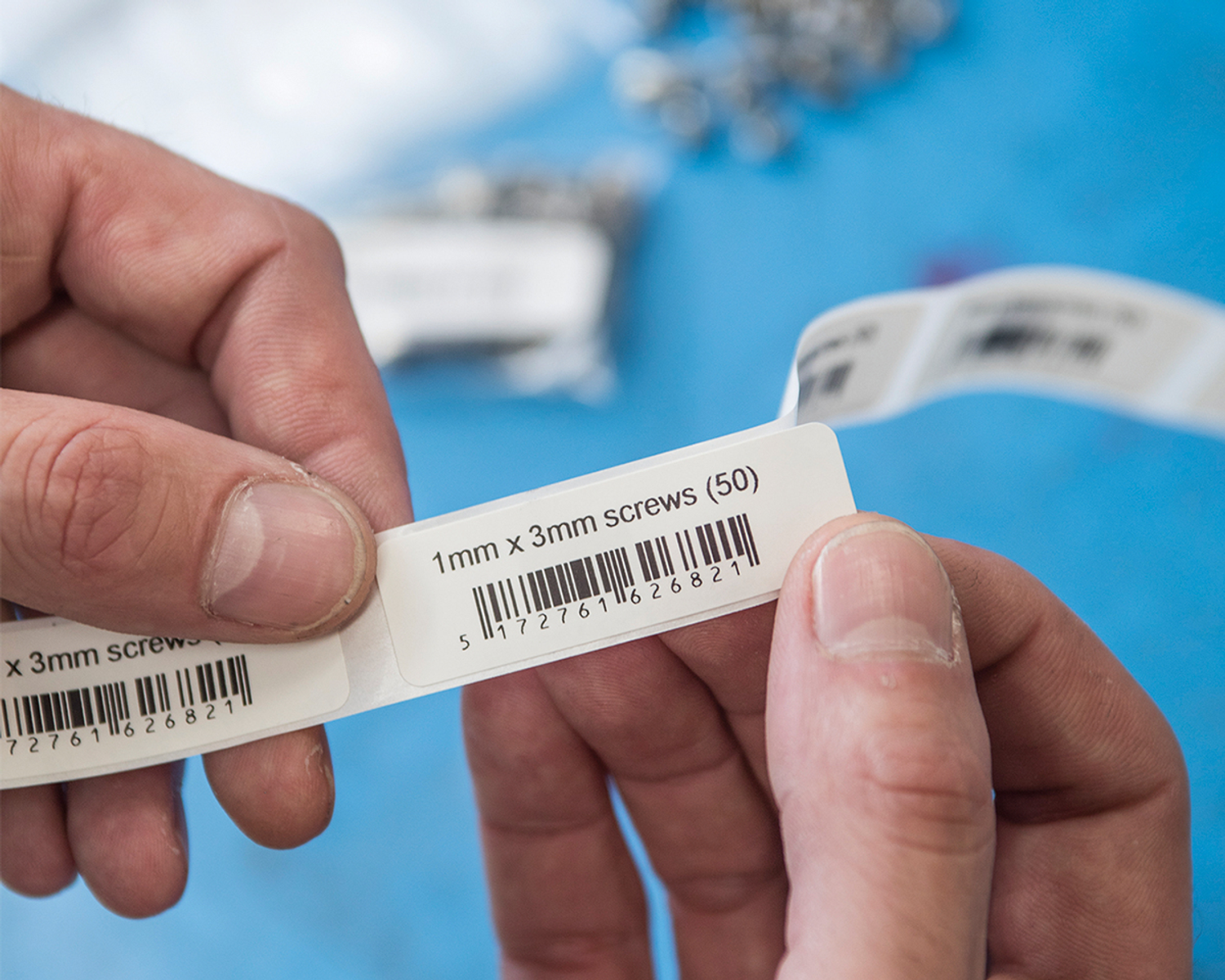 Dłonie trzymające etykiety z tekstem i kodami kreskowymi na niebieskiej powierzchni.