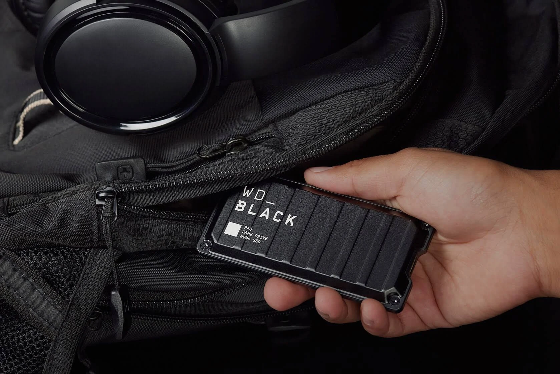 Dłoń trzymająca czarny zewnętrzny dysk twardy obok czarnego plecaka.
