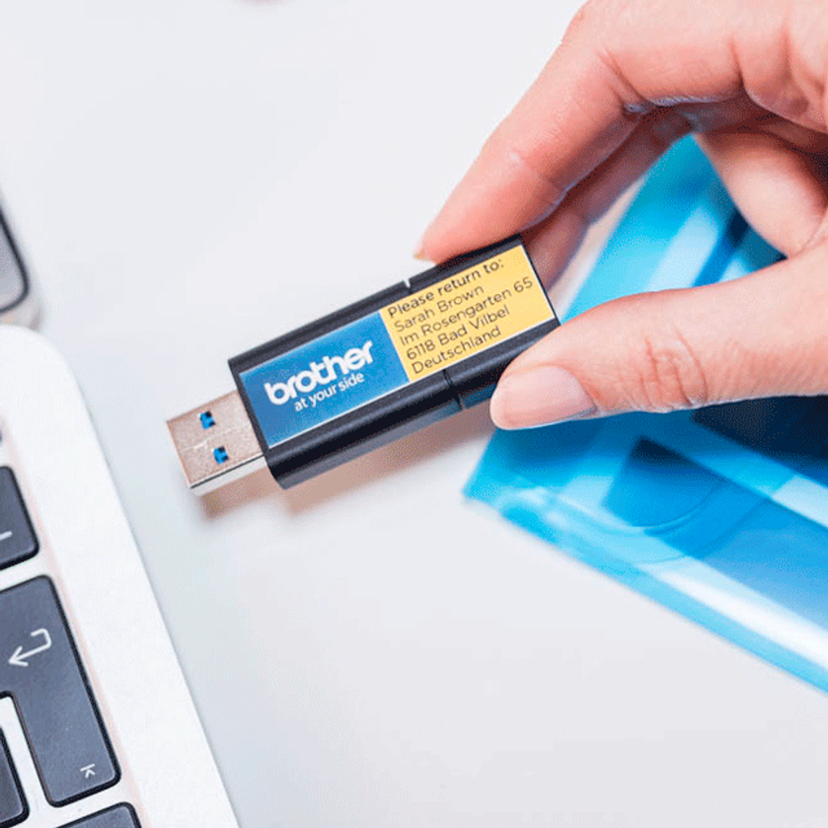 Hand hält USB-Stick mit 'brother'-Label. Der Stick hat ein Rücksendeetikett. Blauer Hintergrund.