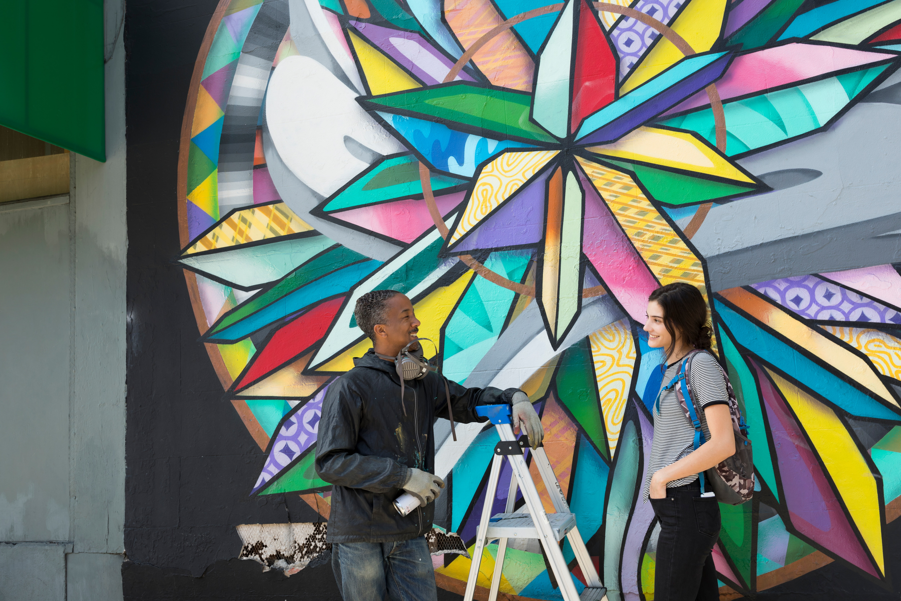 Dwie osoby przy ścianie z graffiti. Jedna trzyma puszkę z farbą, kobieta.