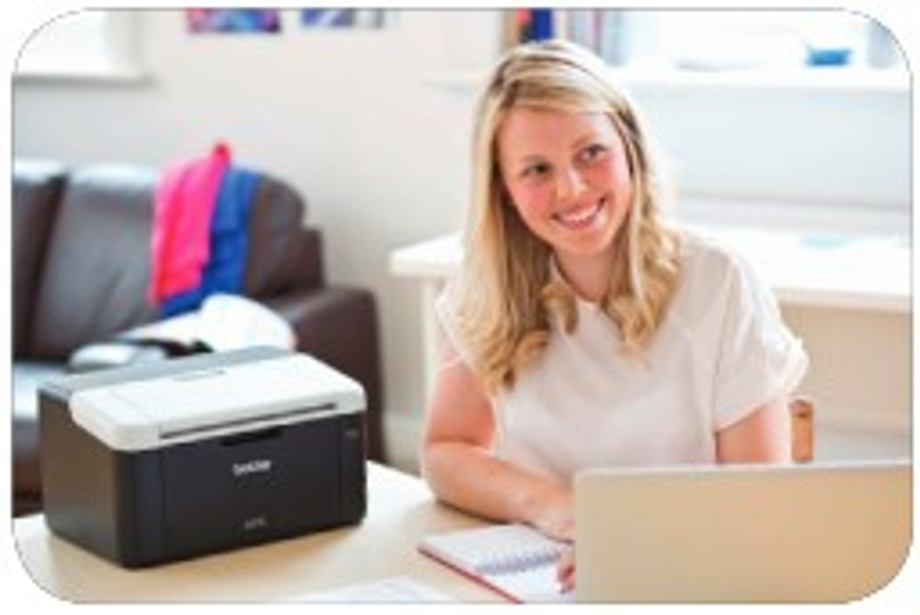 Mujer sonriendo a una laptop. Una impresora y un cuaderno están en la mesa. Un sofá está en el fondo.