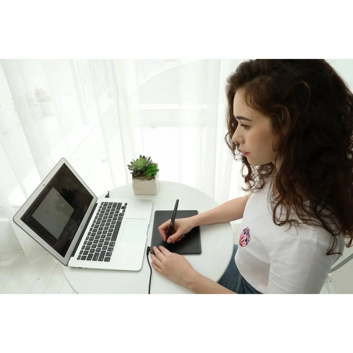 Kobieta używa laptopa i tabletu graficznego na białym stole. Roślina w tle.