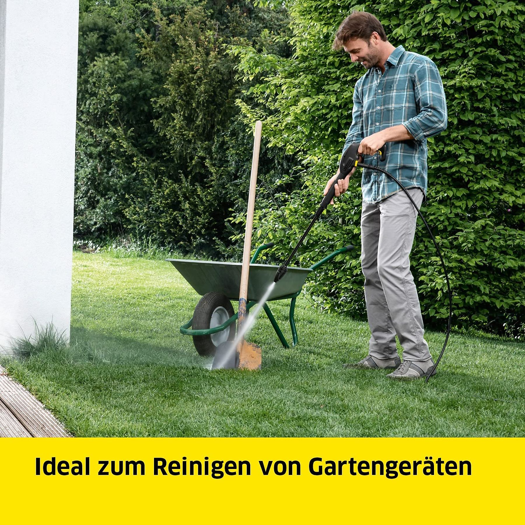 Ein Mann reinigt mit einem Hochdruckreiniger eine Schubkarre auf einem Rasen.