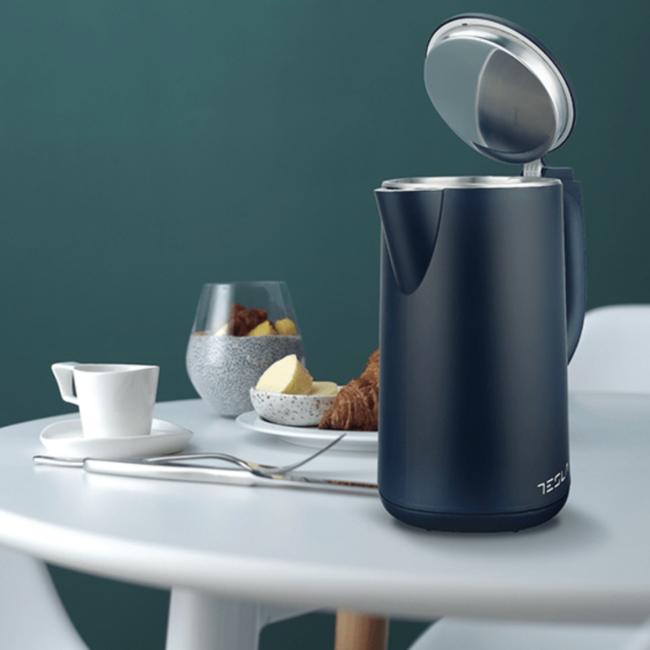Hervidor eléctrico azul oscuro con tapa abierta junto a alimentos de desayuno en mesa blanca.