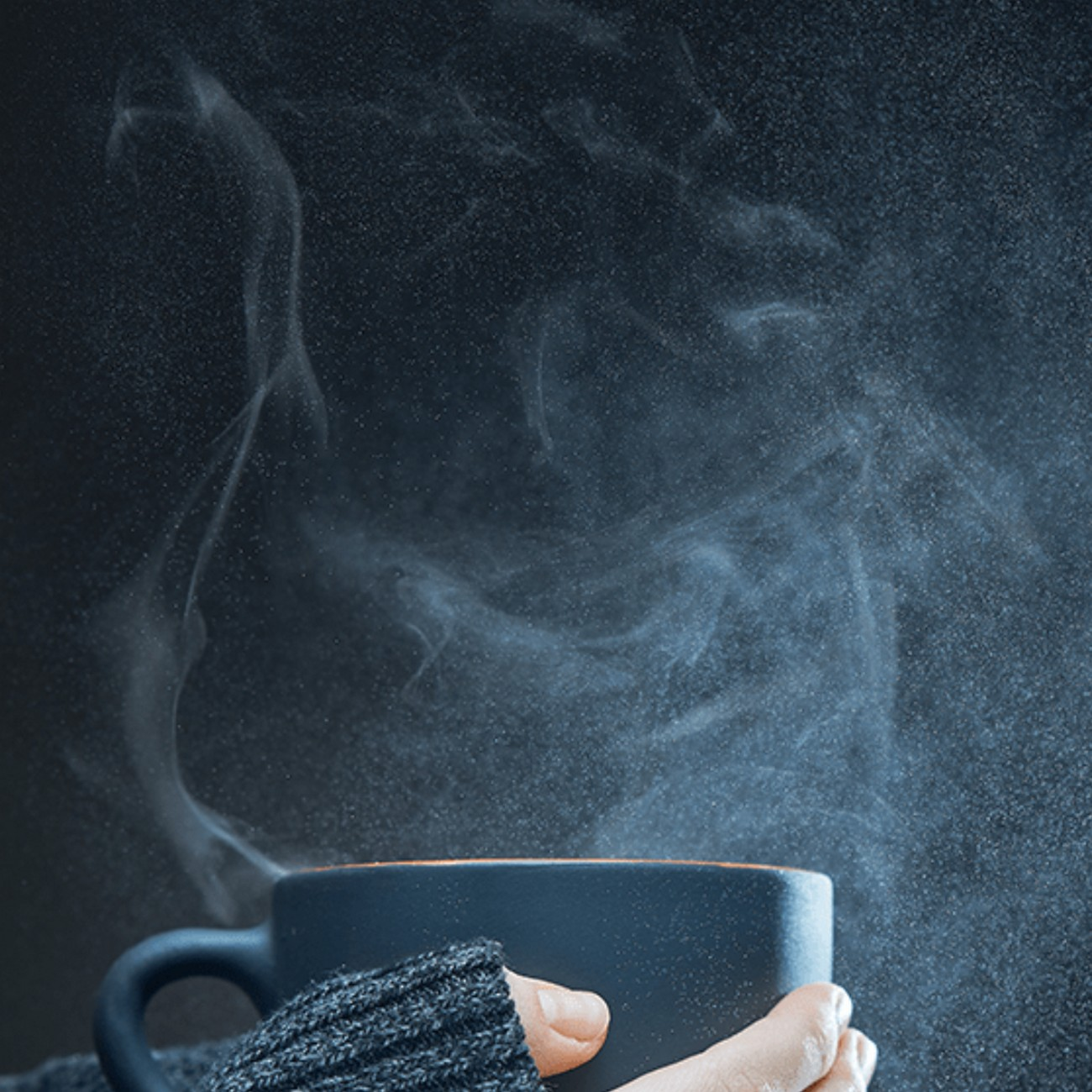 Una taza de café humeante sostenida por una persona con un suéter gris, fondo oscuro.