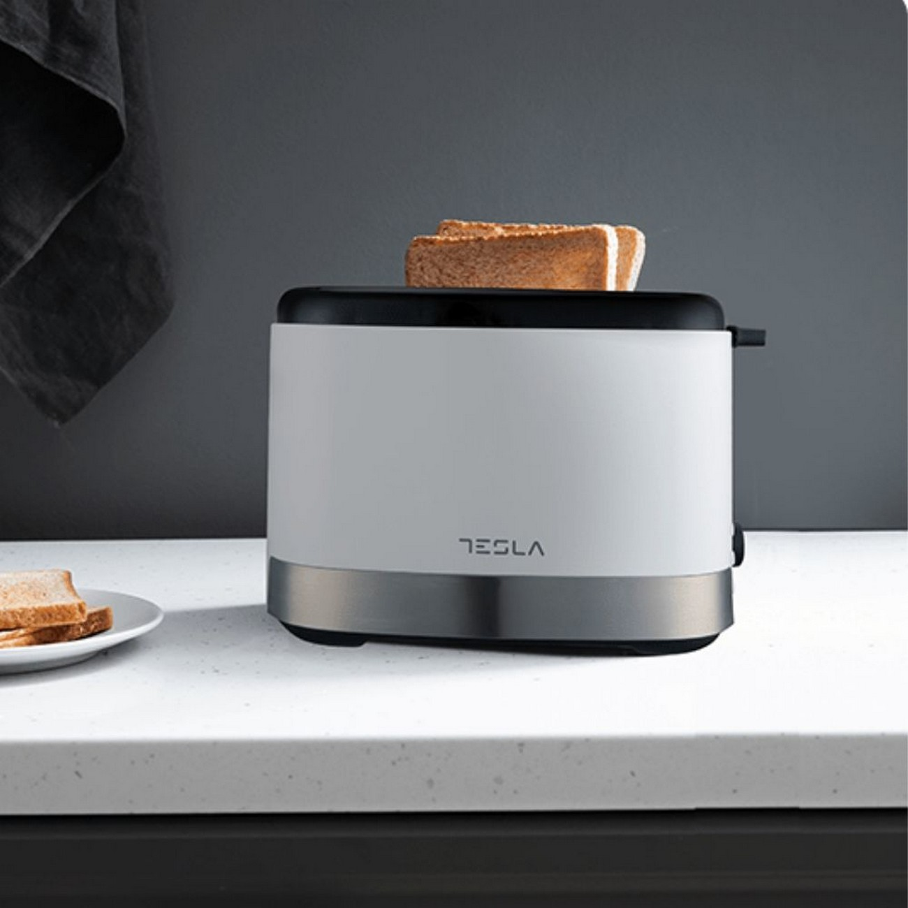 Biały toster Tesla z dwoma kromkami tostów, na białym blacie, na ciemnym tle.