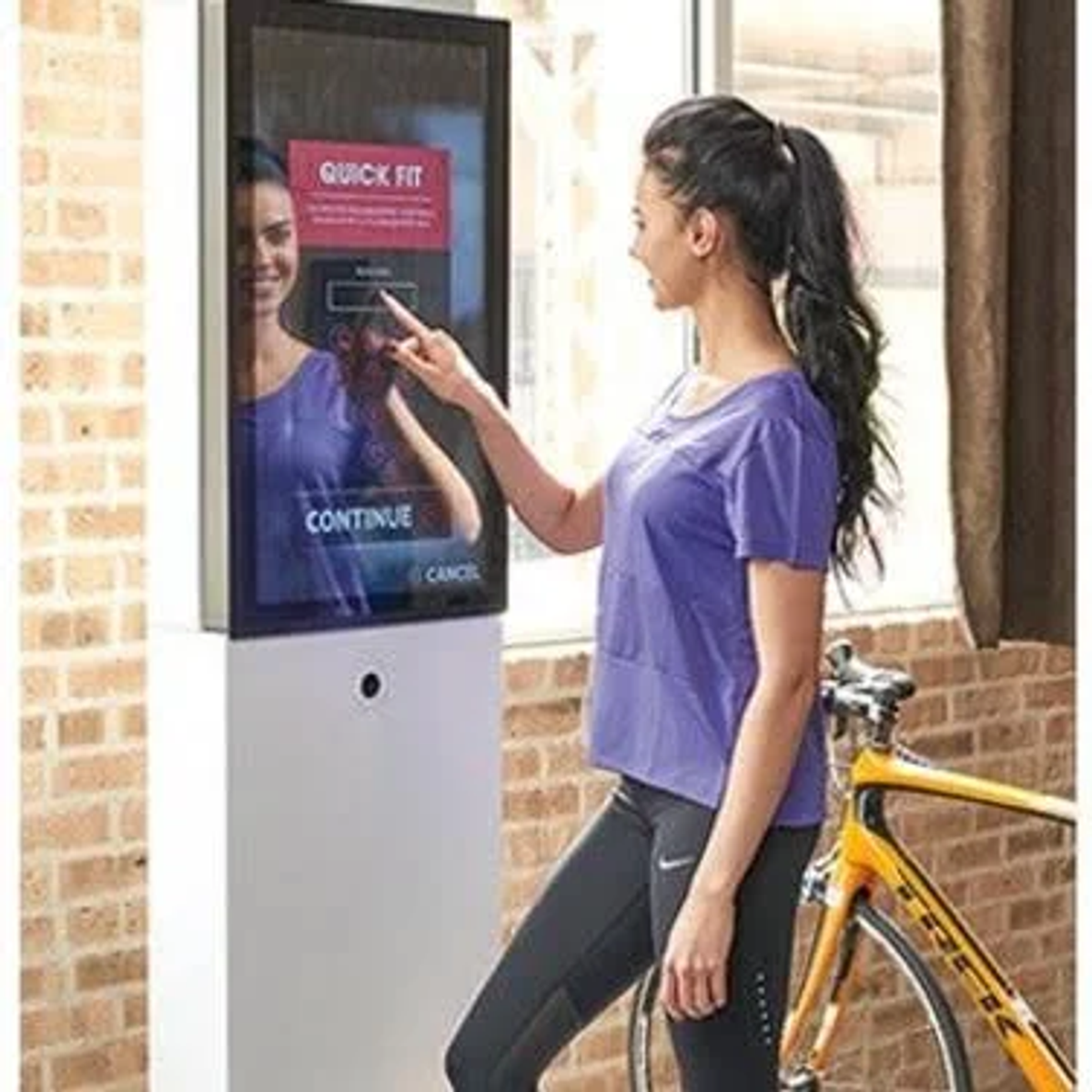 Eine Frau interagiert mit einem Touchscreen. Ein Fahrrad ist in der Nähe. Ein Spiegelbild der Frau ist sichtbar.