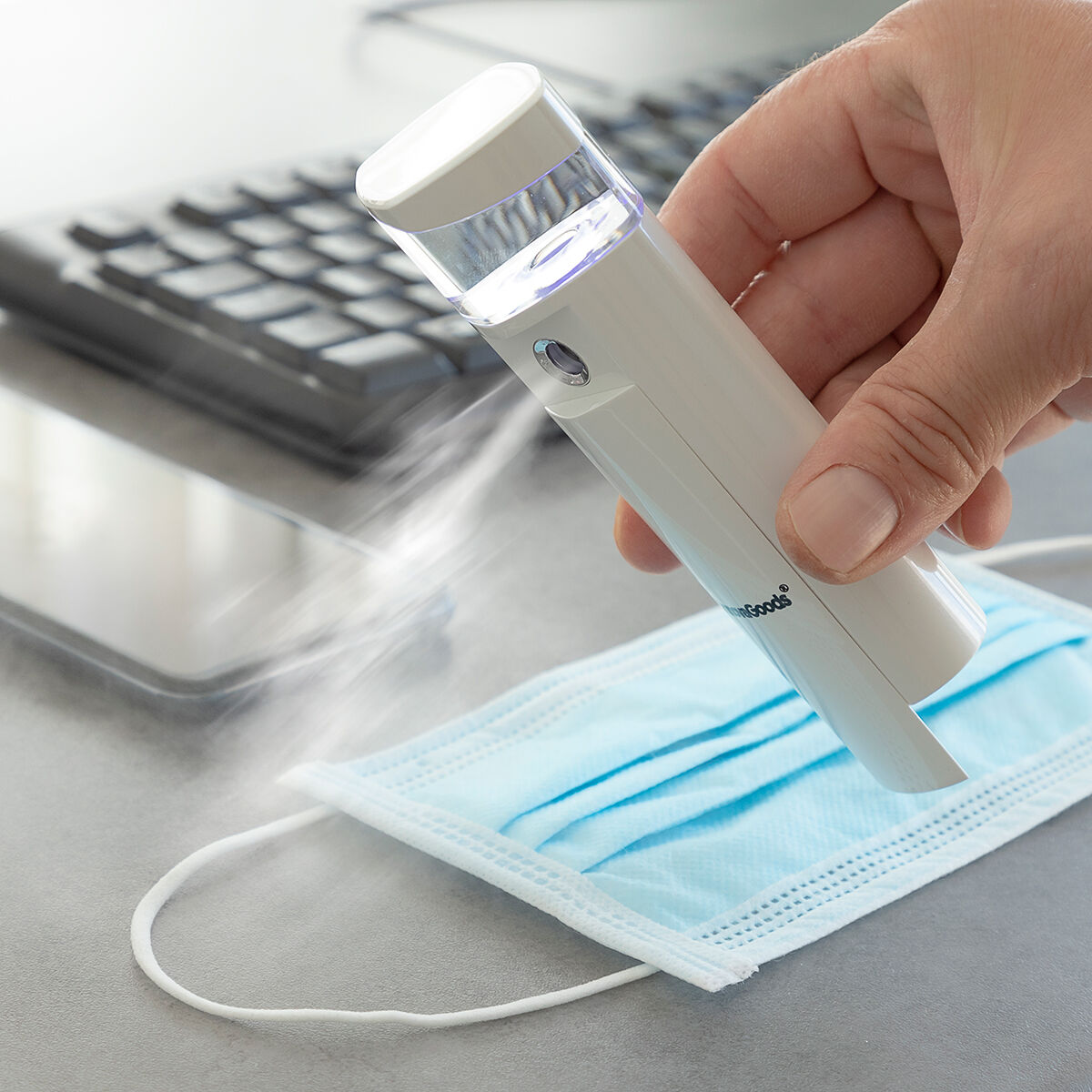 Una mano spruzza un disinfettante bianco su una mascherina blu. Una tastiera e un telefono sono sfocati sullo sfondo.