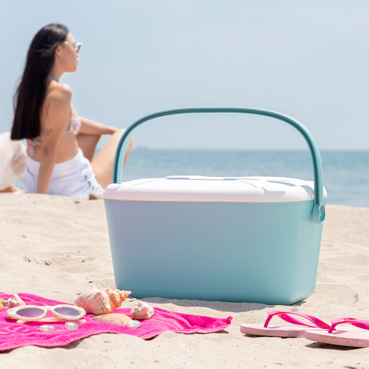 Frau am Strand mit Kühler, Sonnenbrille und rosa Handtuch im Sand.