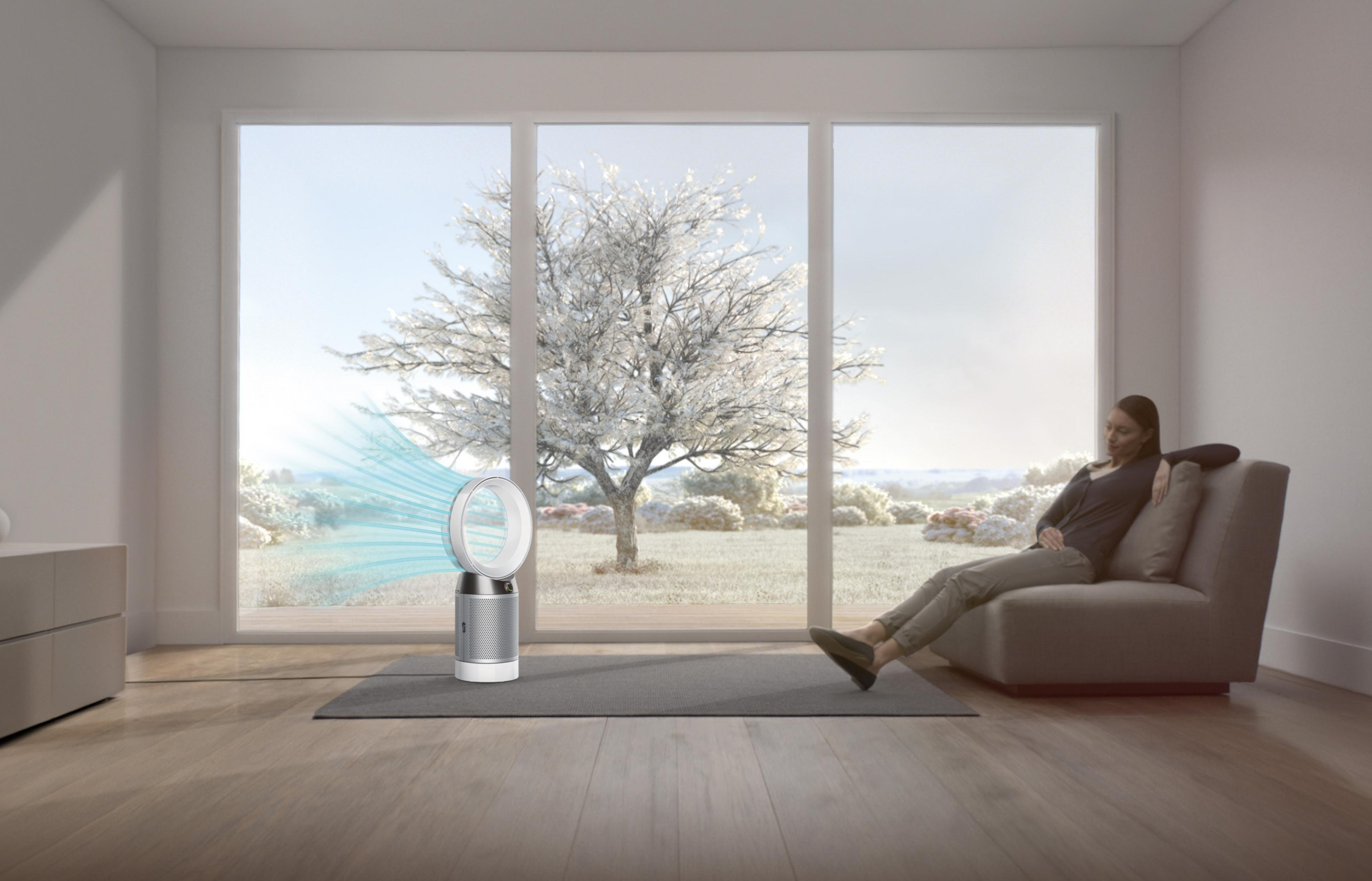 Una mujer sentada cerca de un ventilador Dyson junto a una ventana con paisaje nevado.