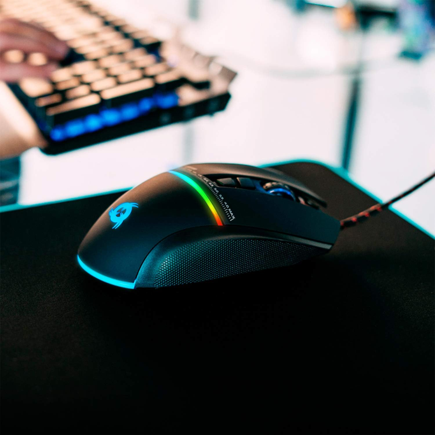 Eine schwarze Gaming-Maus mit einem mehrfarbigen Lichtstreifen, auf einem schwarzen Mauspad, mit Tastaturtasten im Hintergrund.