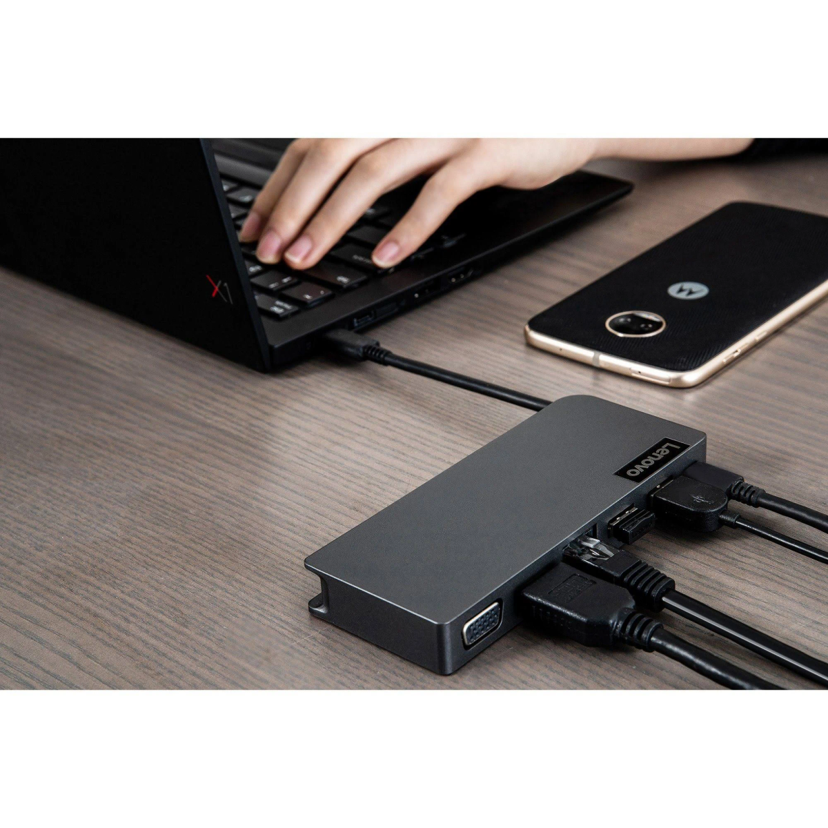 Le mani di una persona che scrive su un laptop nero vicino a un hub Lenovo con cavi collegati e un telefono.